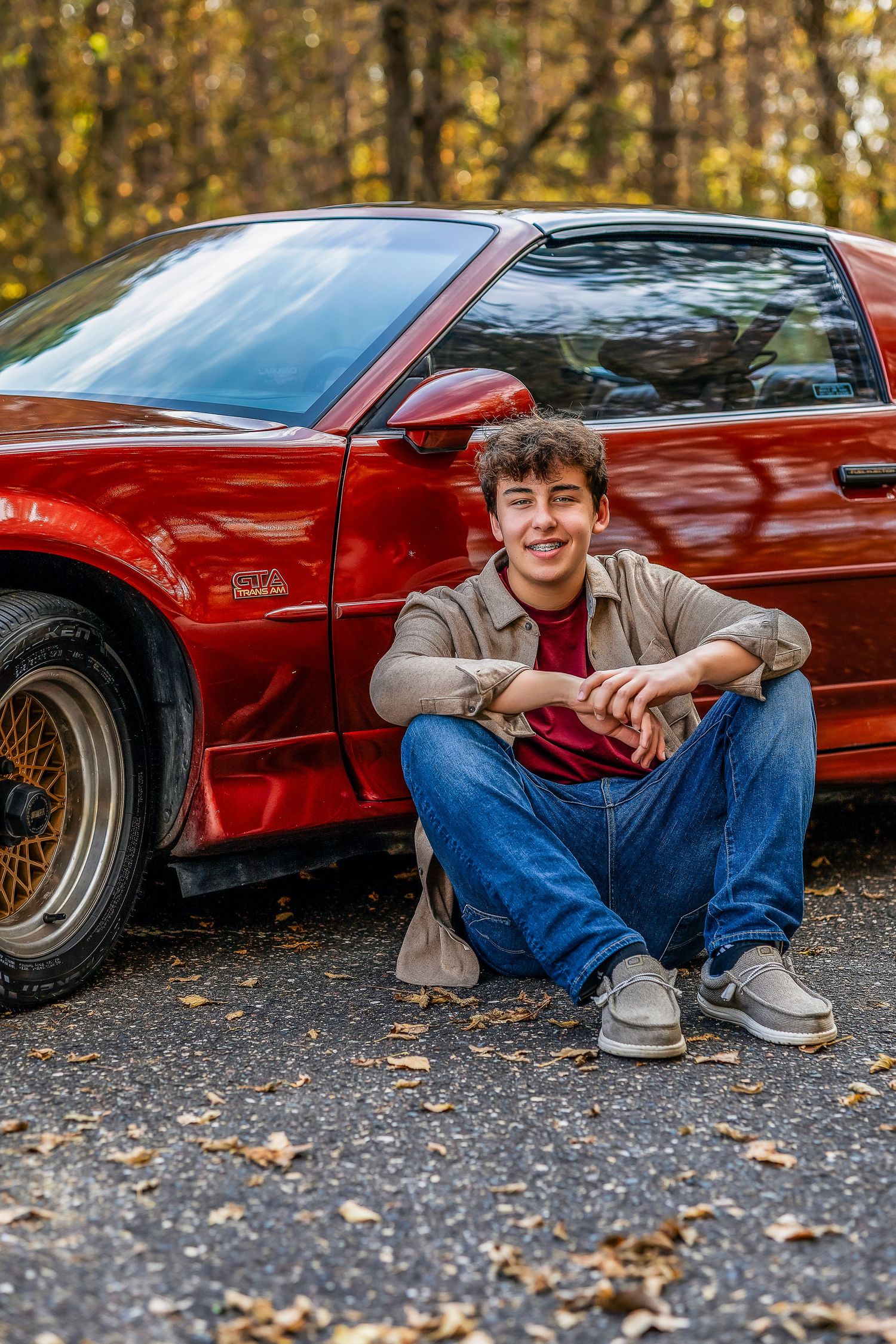 Young man sitting on the ground wearing jeans next to a red car