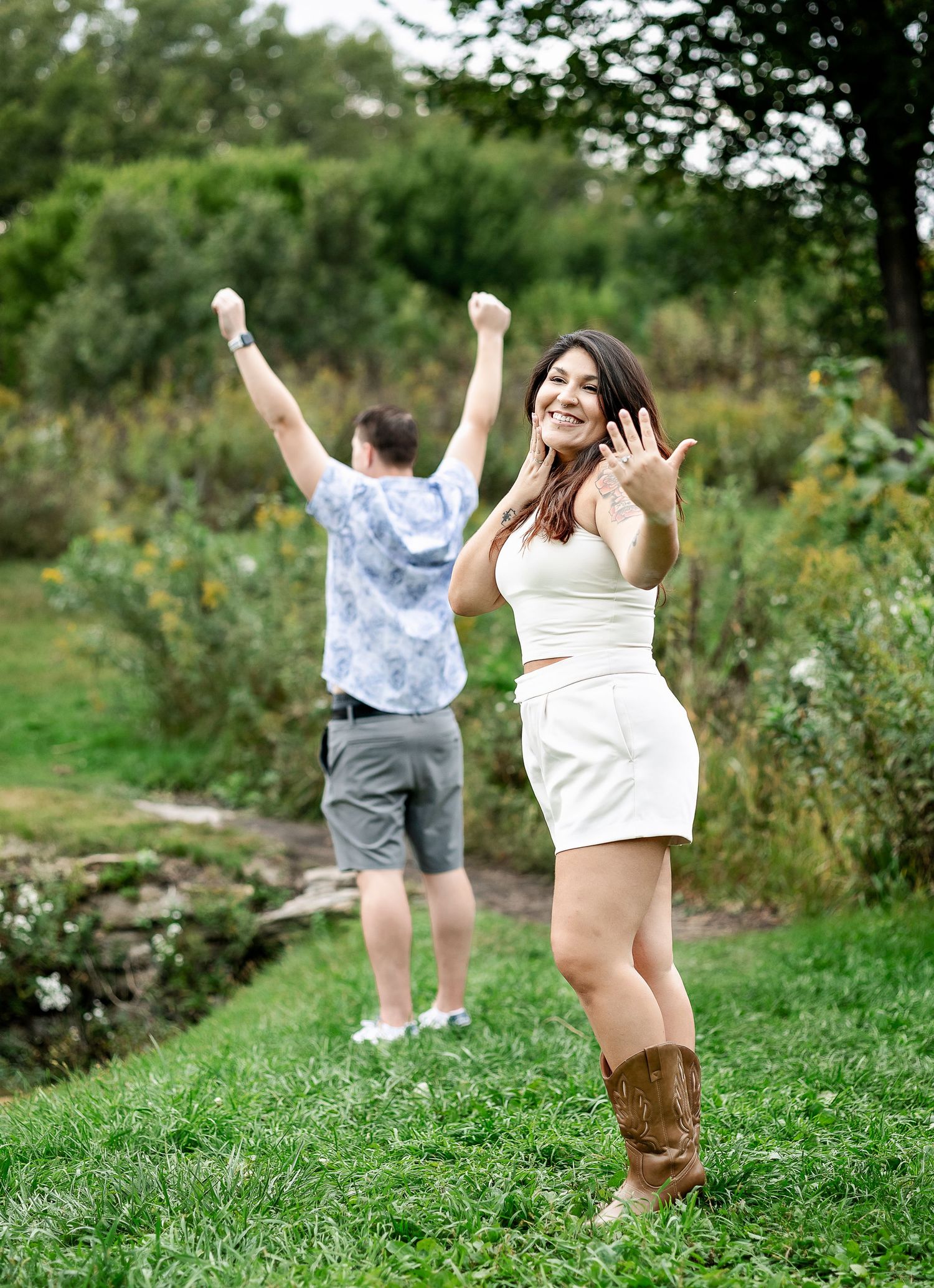 Girl holding engagement ring up in the air while man celebrating behind her