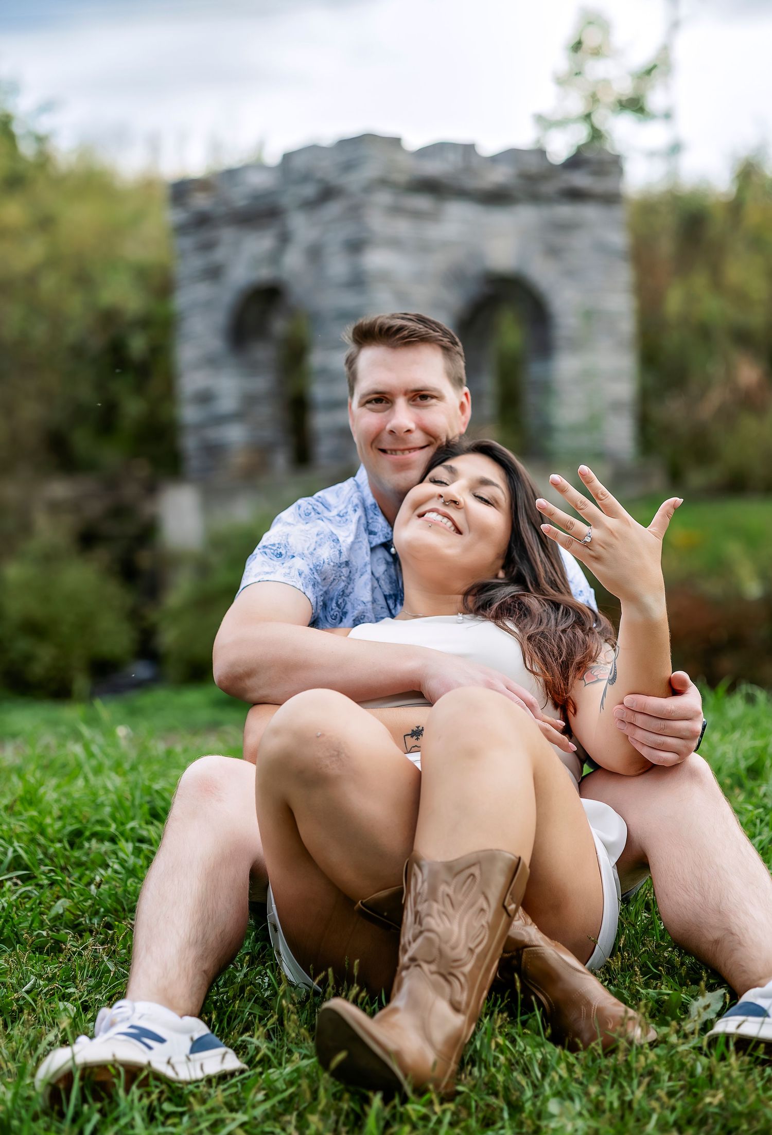 Man and woman cuddling in the grass holding up engagement ring