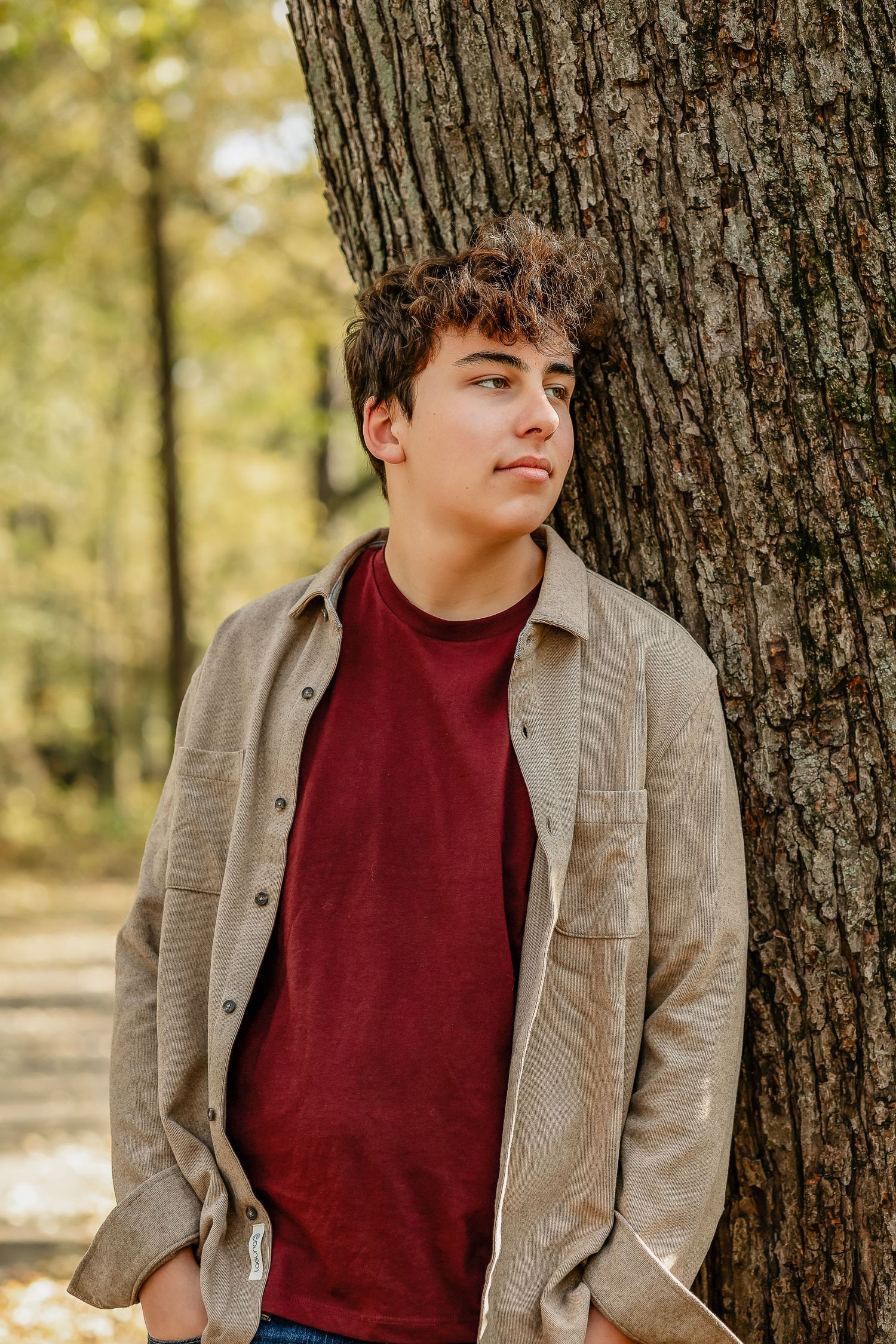 Young man wearing a red shirt looking out in the distance, leaning against a tree.