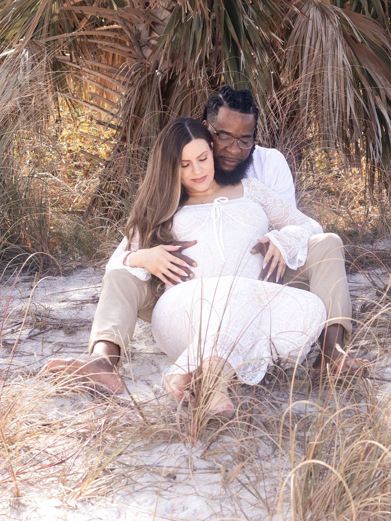 Couple sharing a tender maternity moment on sandy beach, surrounded by palm trees and dune grass.