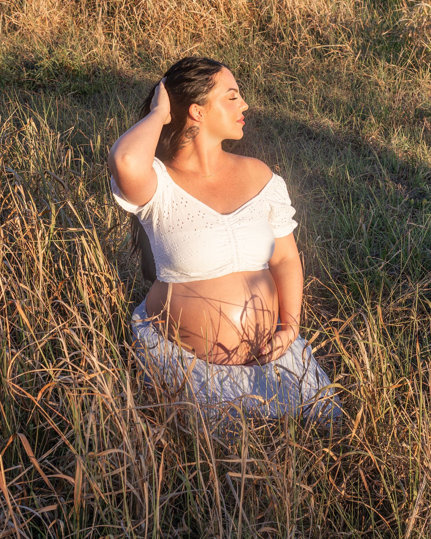 Pregnant woman in white crop top and skirt sitting in golden grass field during sunset.