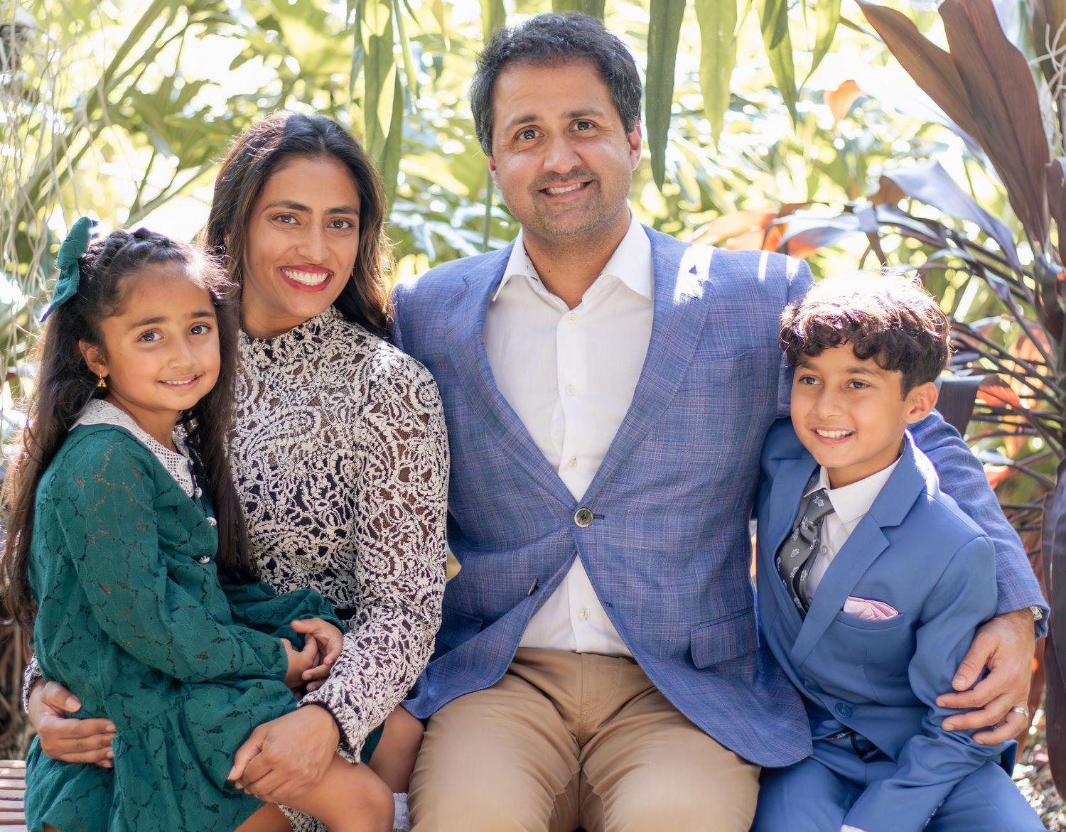 A smiling family of four poses outdoors, with two adults and two children dressed in elegant attire.