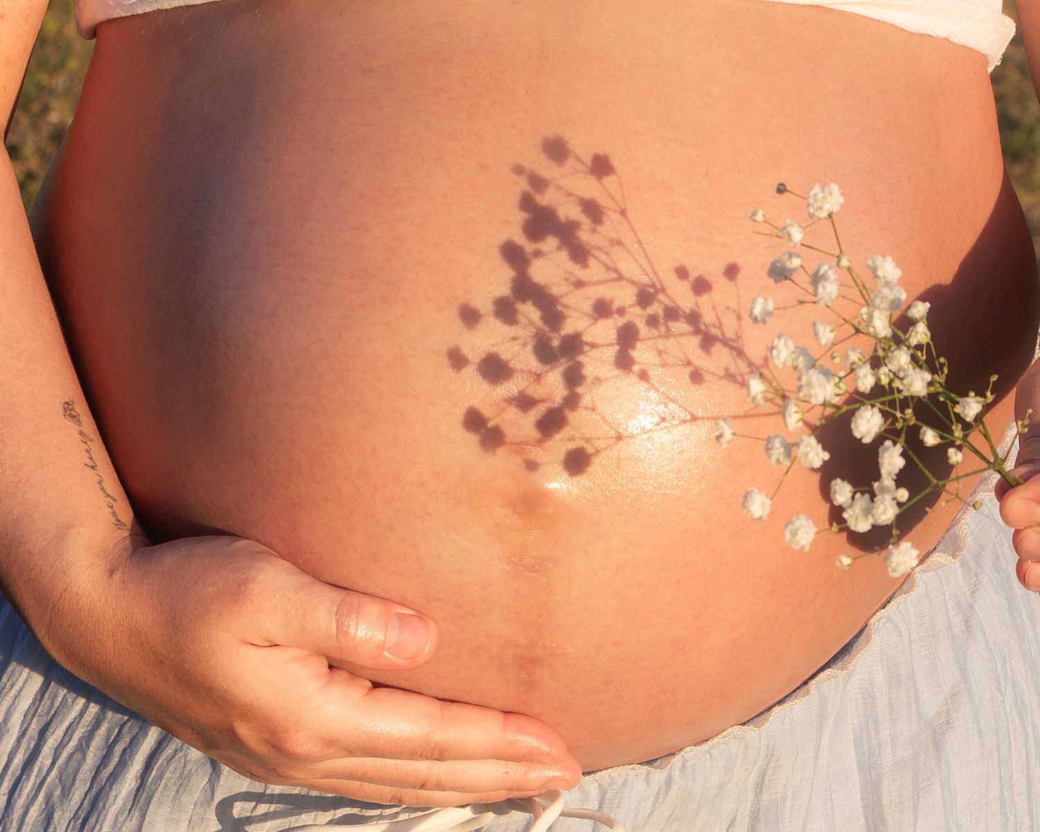 Close-up of a pregnant woman's bare belly adorned with delicate dried wildflowers in warm golden sunlight.