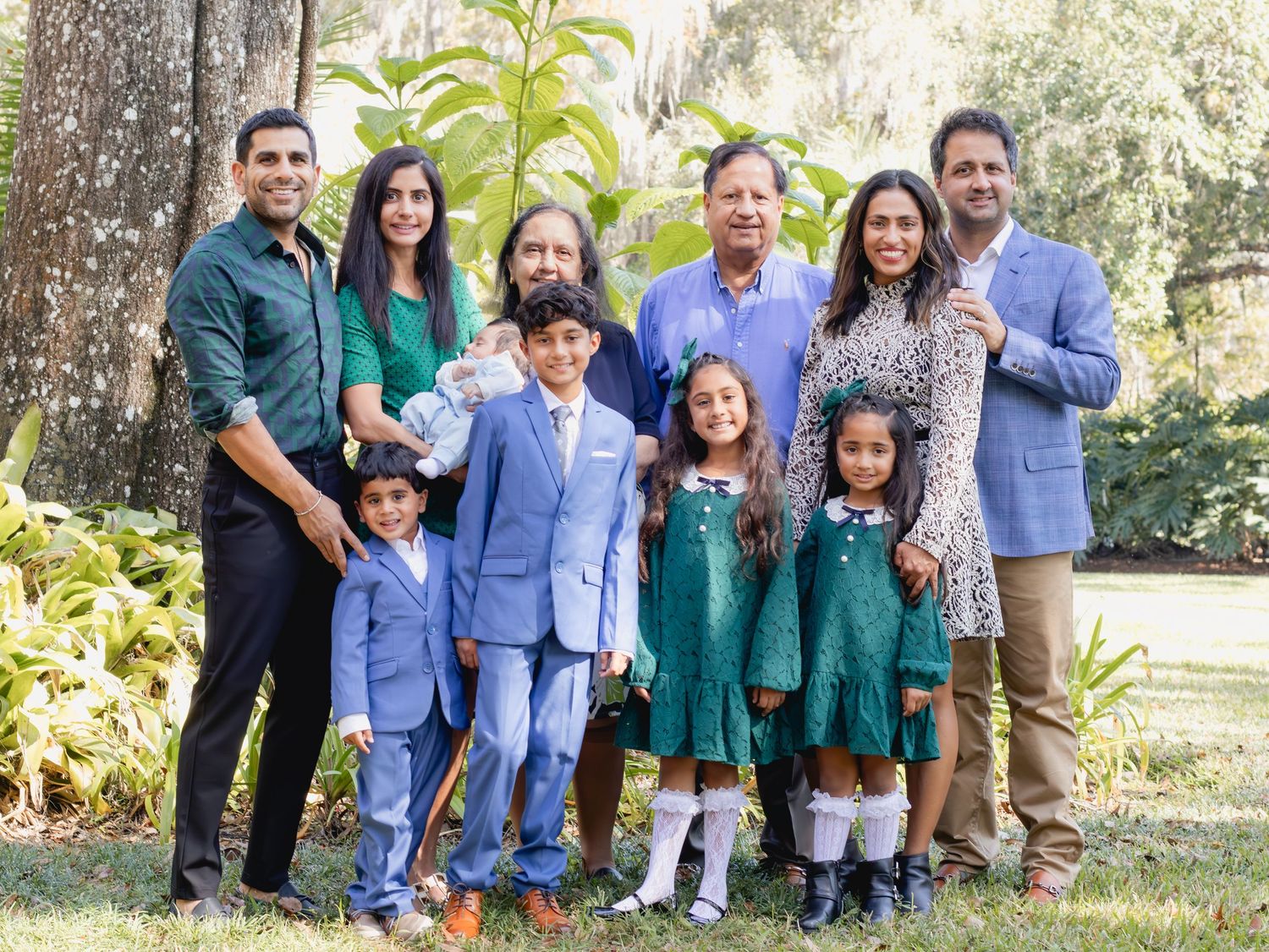 Large multigenerational South Asian family posing together outdoors in a lush green park setting for a portrait.