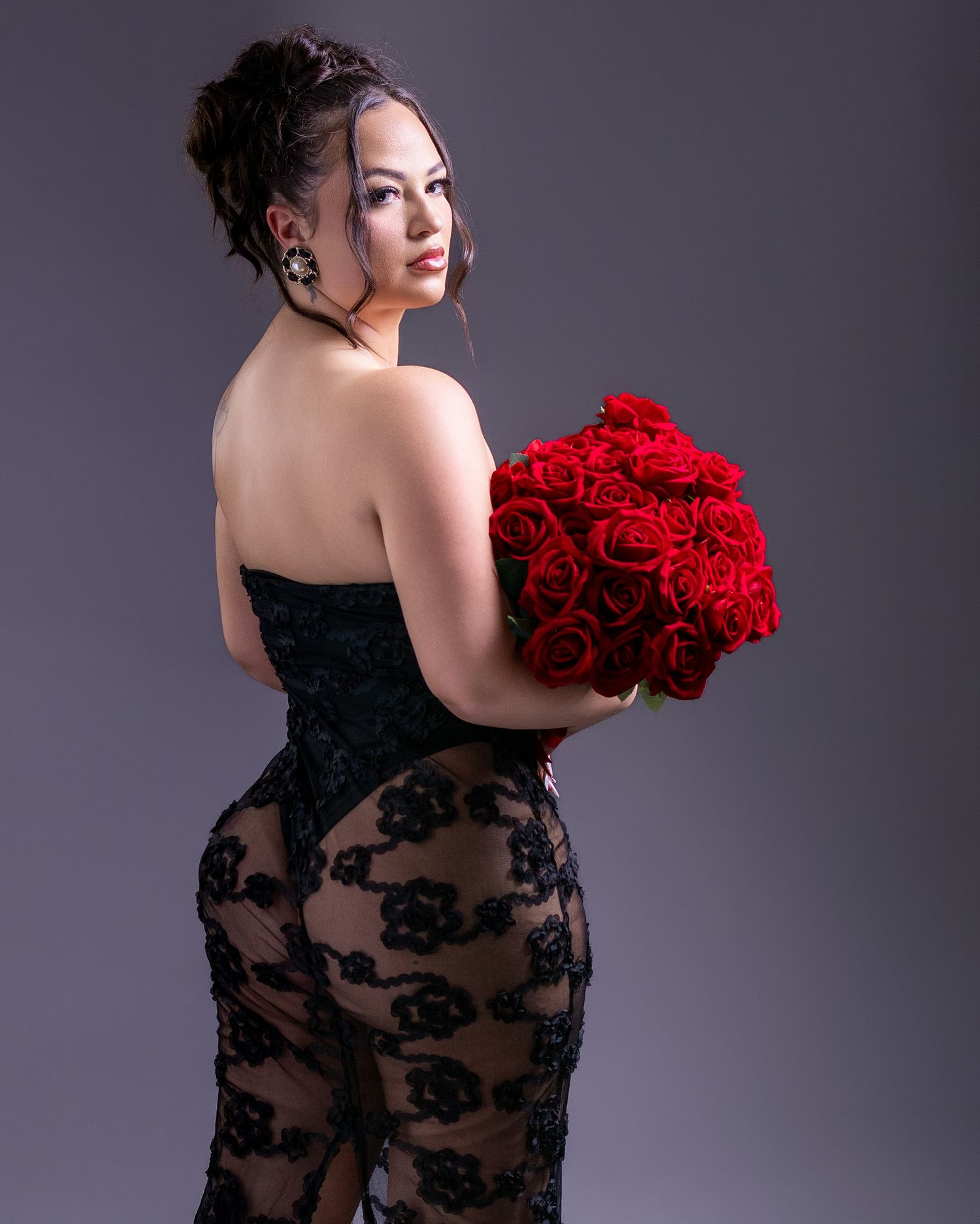 Woman in black lace gown holding red roses bouquet, posing elegantly in studio portrait photography.