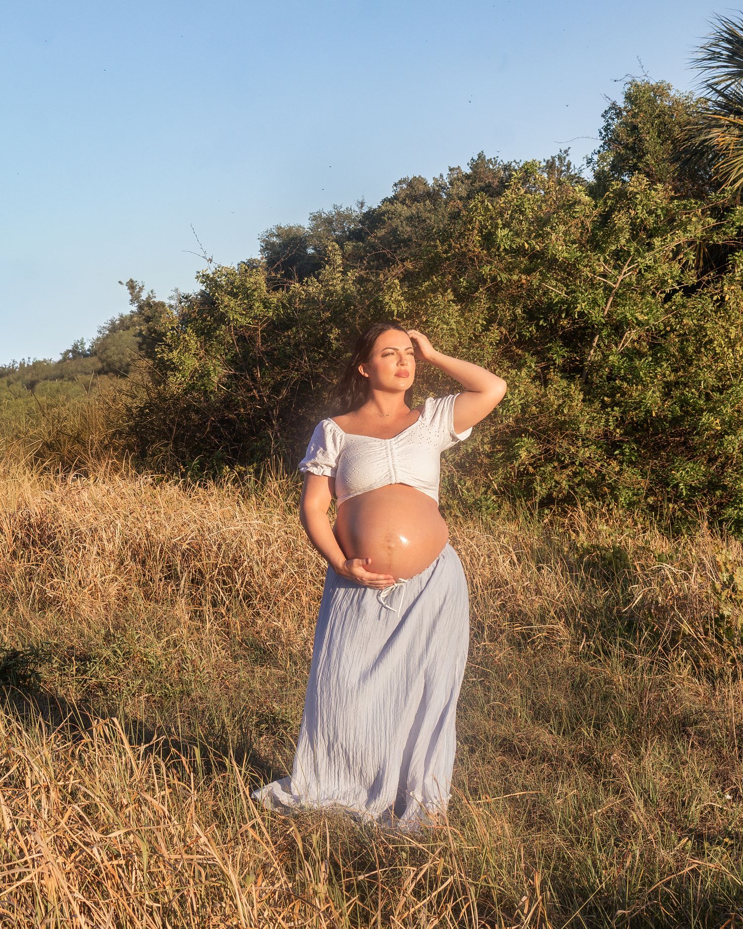 Pregnant woman in white crop top and blue skirt holding belly in golden hour sunlit field with green trees.