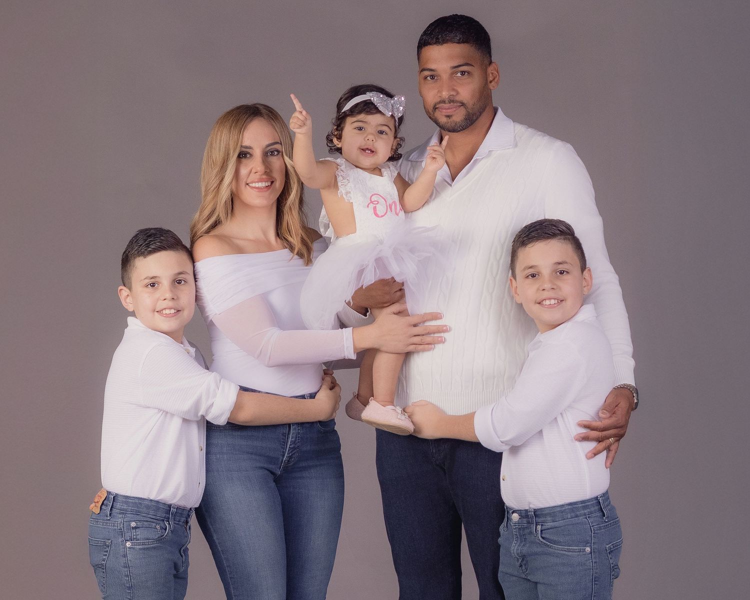 Family of five posing in white tops and jeans for a studio portrait, with parents holding a baby girl in a white dress.