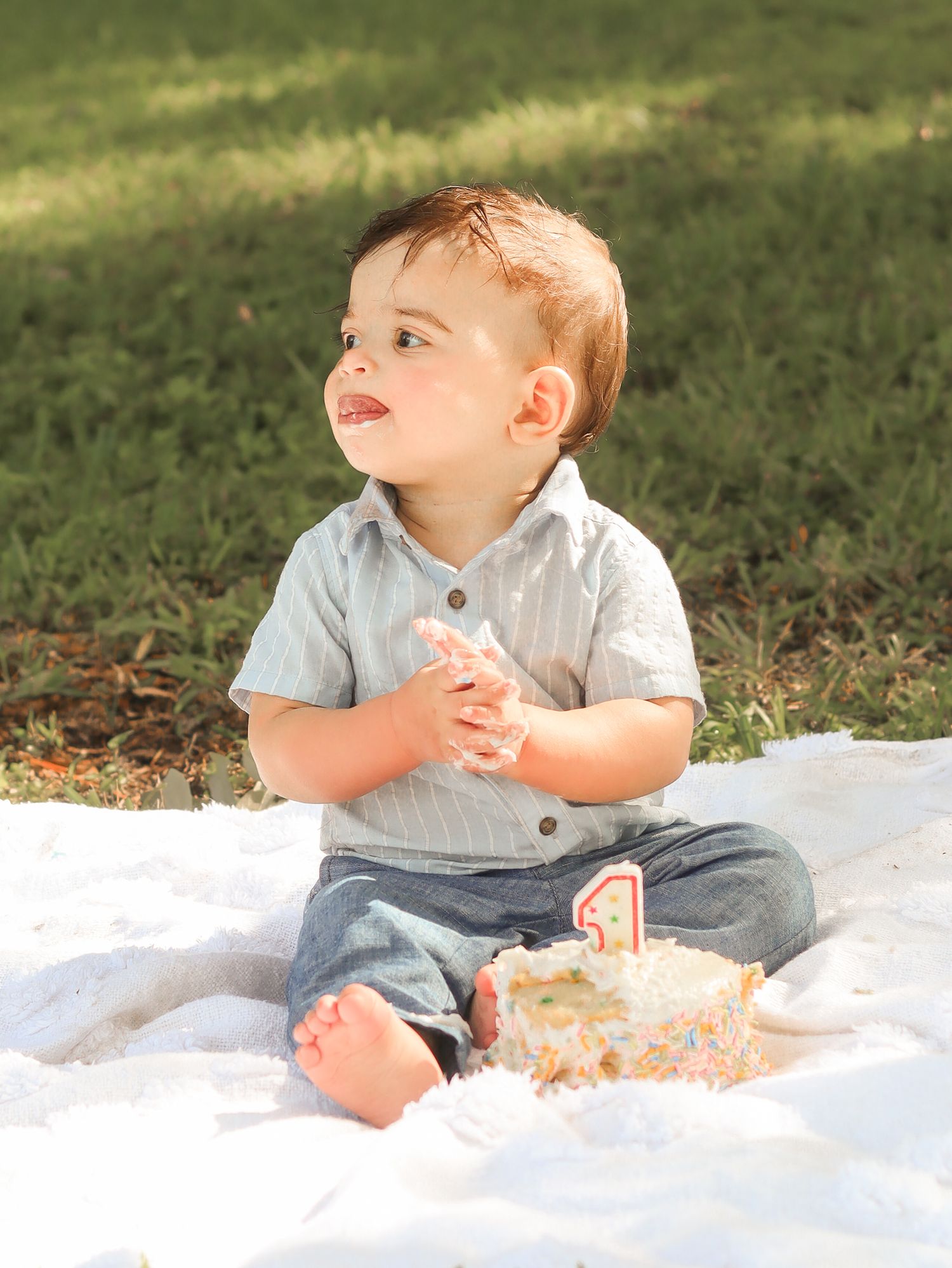 Toddler boy sitting on white blanket outdoors celebrating first birthday with cake, wearing blue shirt and jeans.