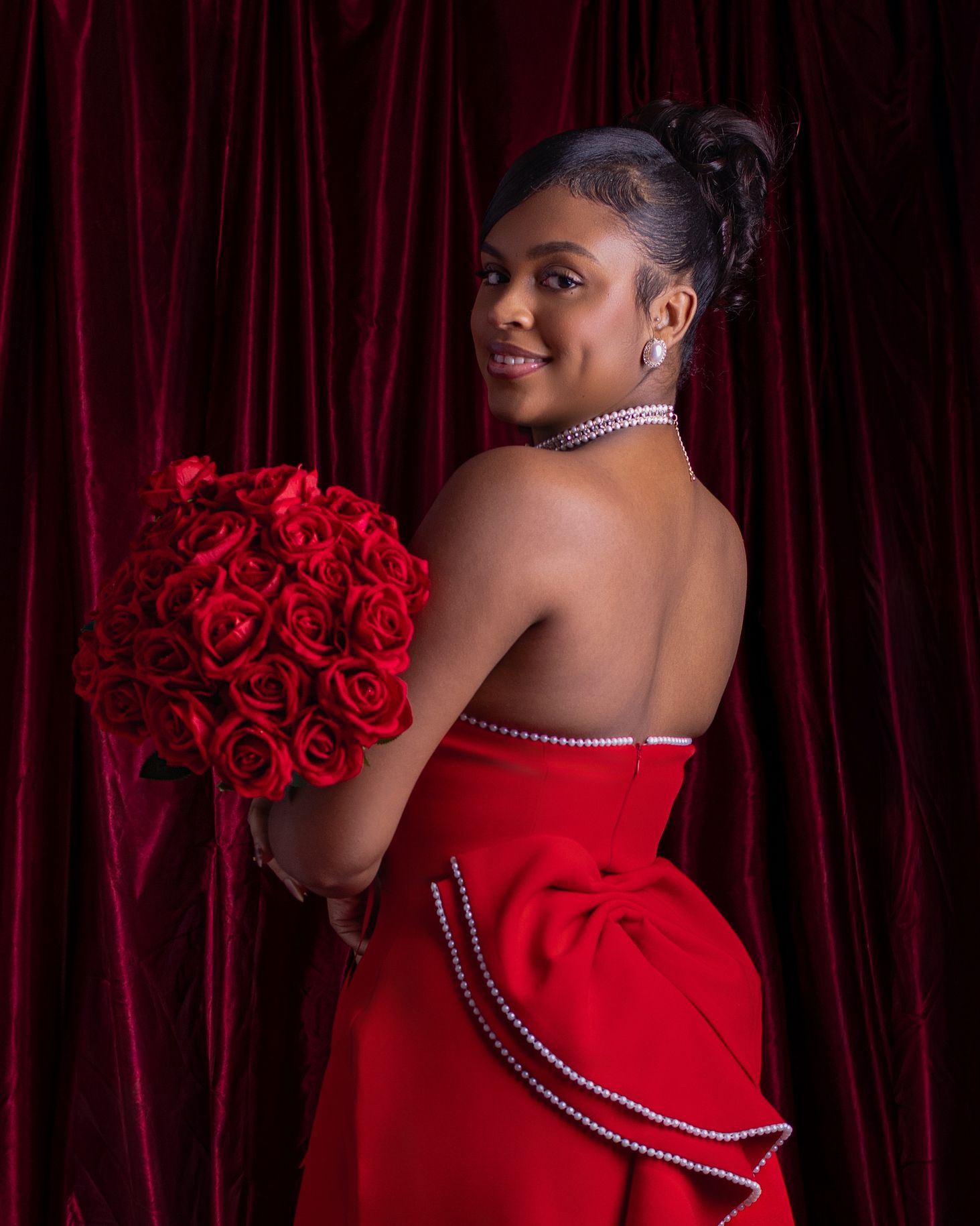 Woman in red strapless gown holding red rose bouquet, pearl jewelry, posing against burgundy velvet curtain backdrop.