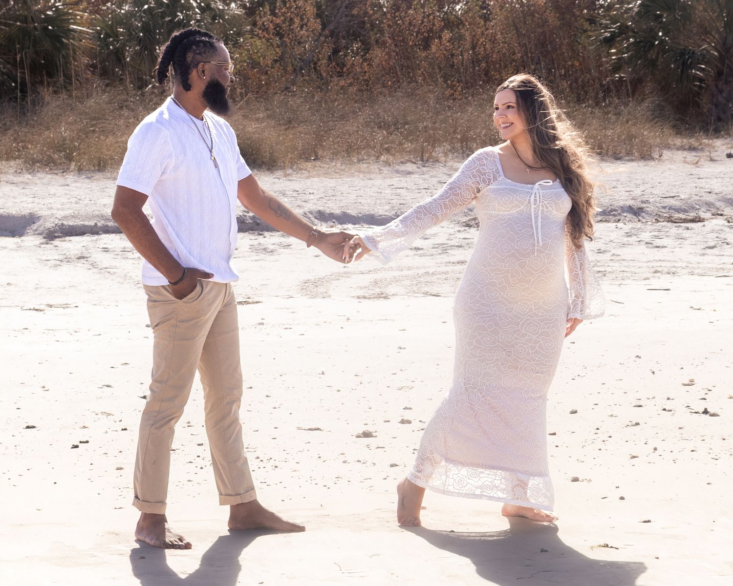 Couple in white outfits holding hands on a sandy beach, woman in lace maternity dress during outdoor photoshoot.