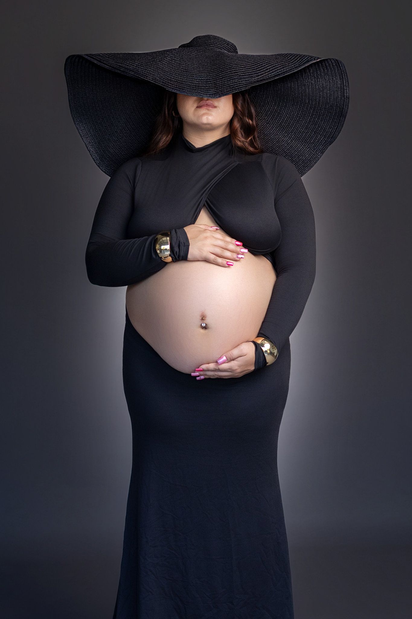 Pregnant woman in black dress and wide-brim hat cradling baby bump, wearing gold bracelets and pink nails.