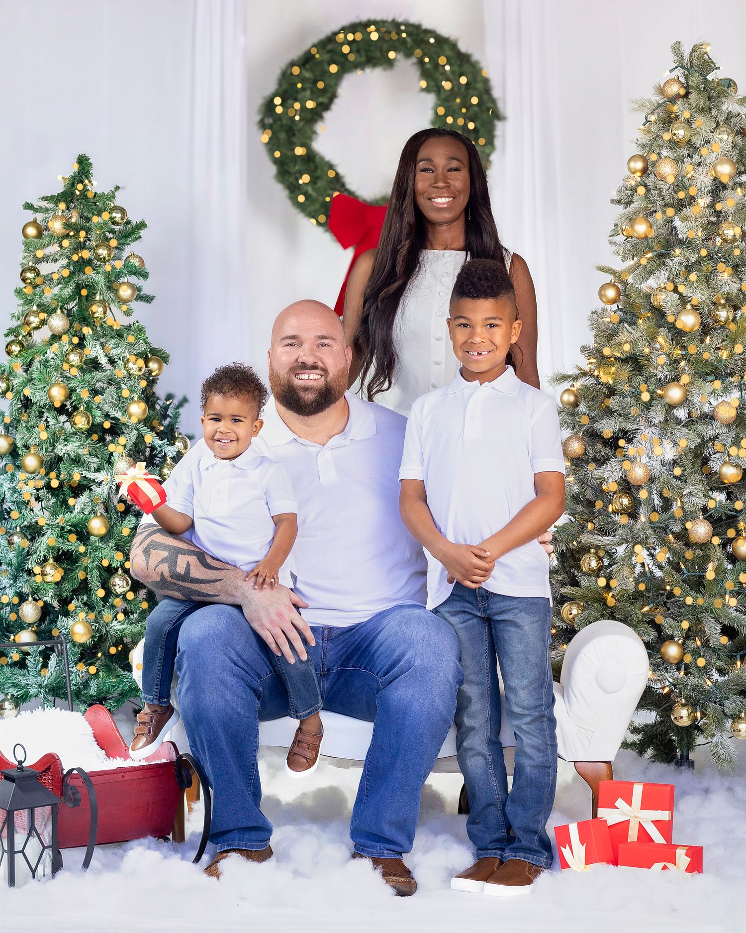 Family of four poses joyfully in front of decorated Christmas trees with gifts and a festive wreath backdrop.