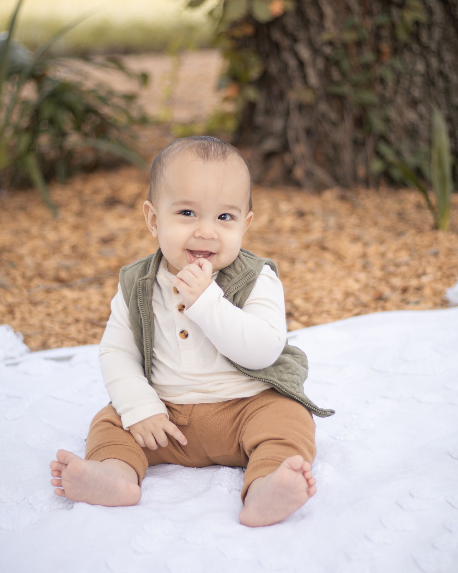 Smiling baby sitting outdoors on white blanket, wearing green vest, white shirt, and brown pants, with fall foliage background.