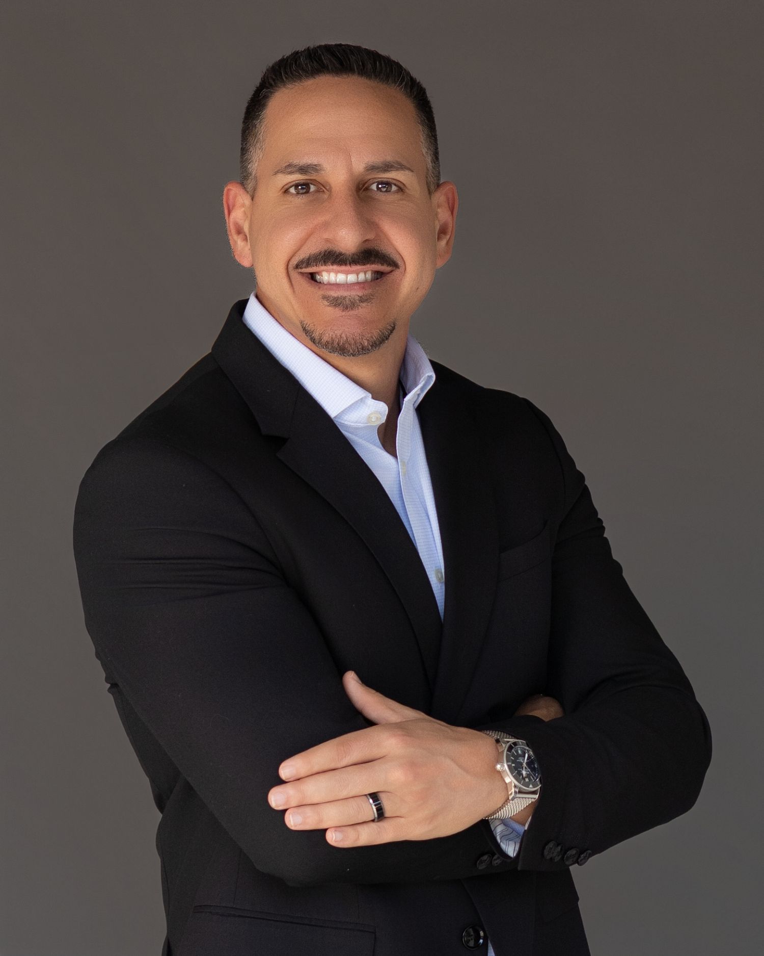 Confident businessman in black suit smiling with arms crossed on gray backdrop.