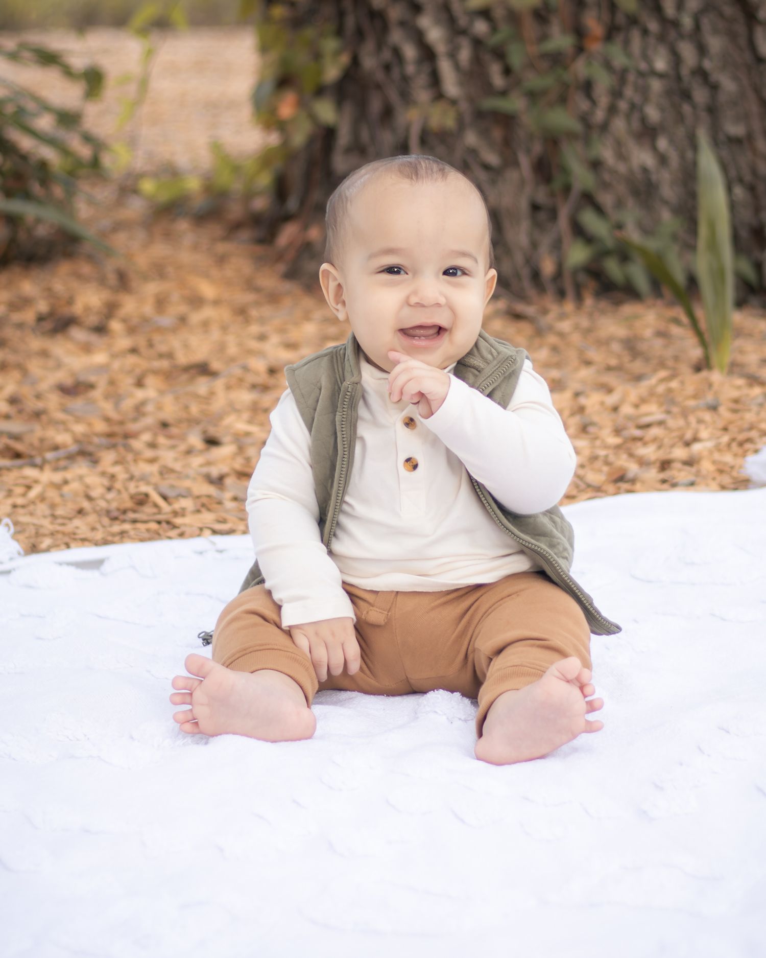 Happy baby boy in green vest sitting outdoors on white blanket near tree.