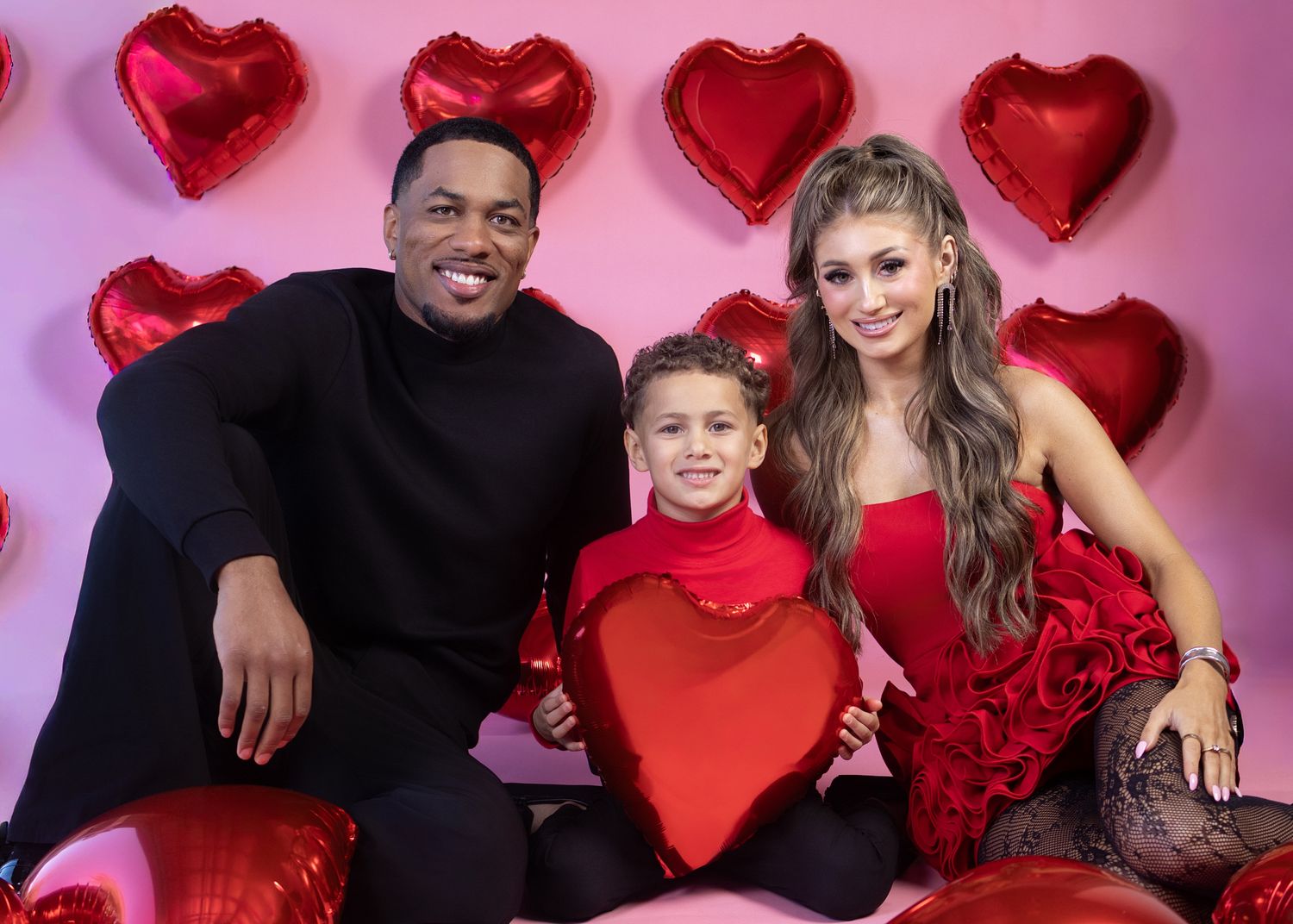 Family of three posing with red heart balloons on a pink Valentine's Day backdrop, wearing festive red outfits.