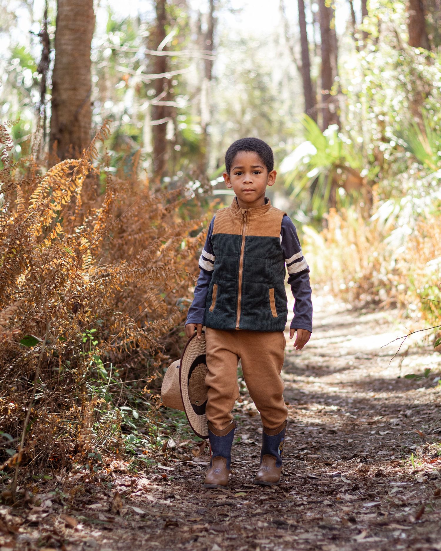 Young boy walking on a forest trail holding a hat, wearing a brown vest and tan pants.