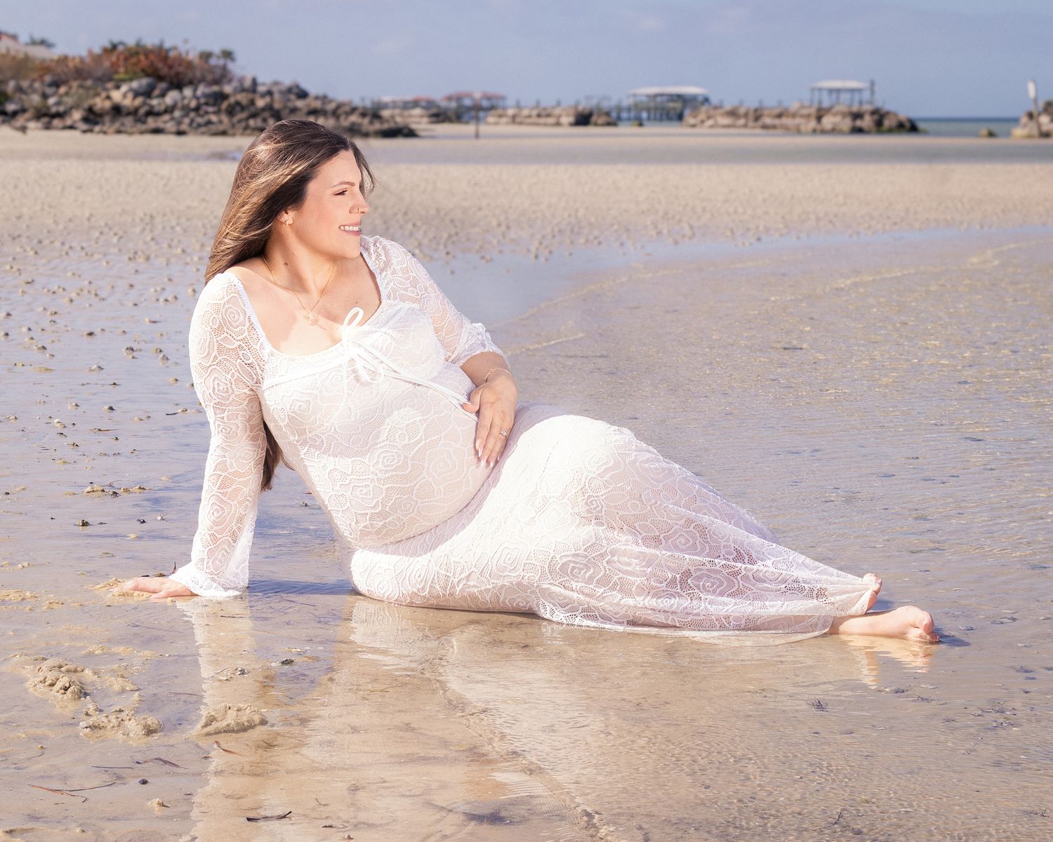 Pregnant woman in white lace gown sitting gracefully on a sandy beach shoreline with calm ocean waters reflecting sunlight.