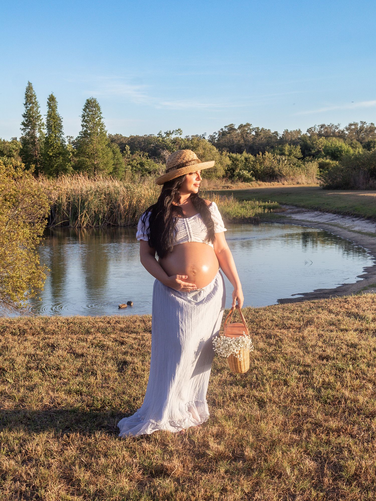 Pregnant woman in white dress and straw hat holding basket by a serene pond in golden hour light.
