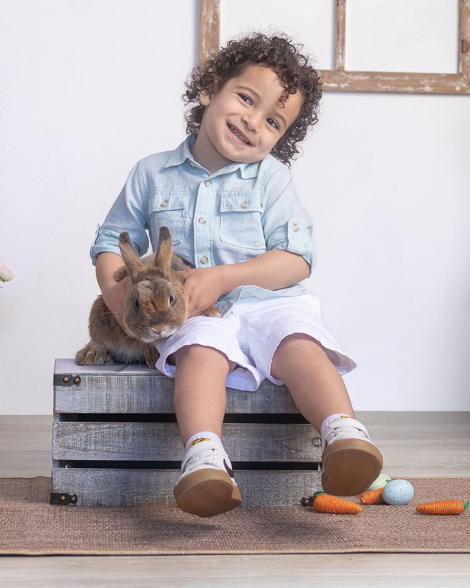Smiling young boy in blue shirt and white shorts sitting on wooden crate holding a brown bunny with Easter eggs nearby.