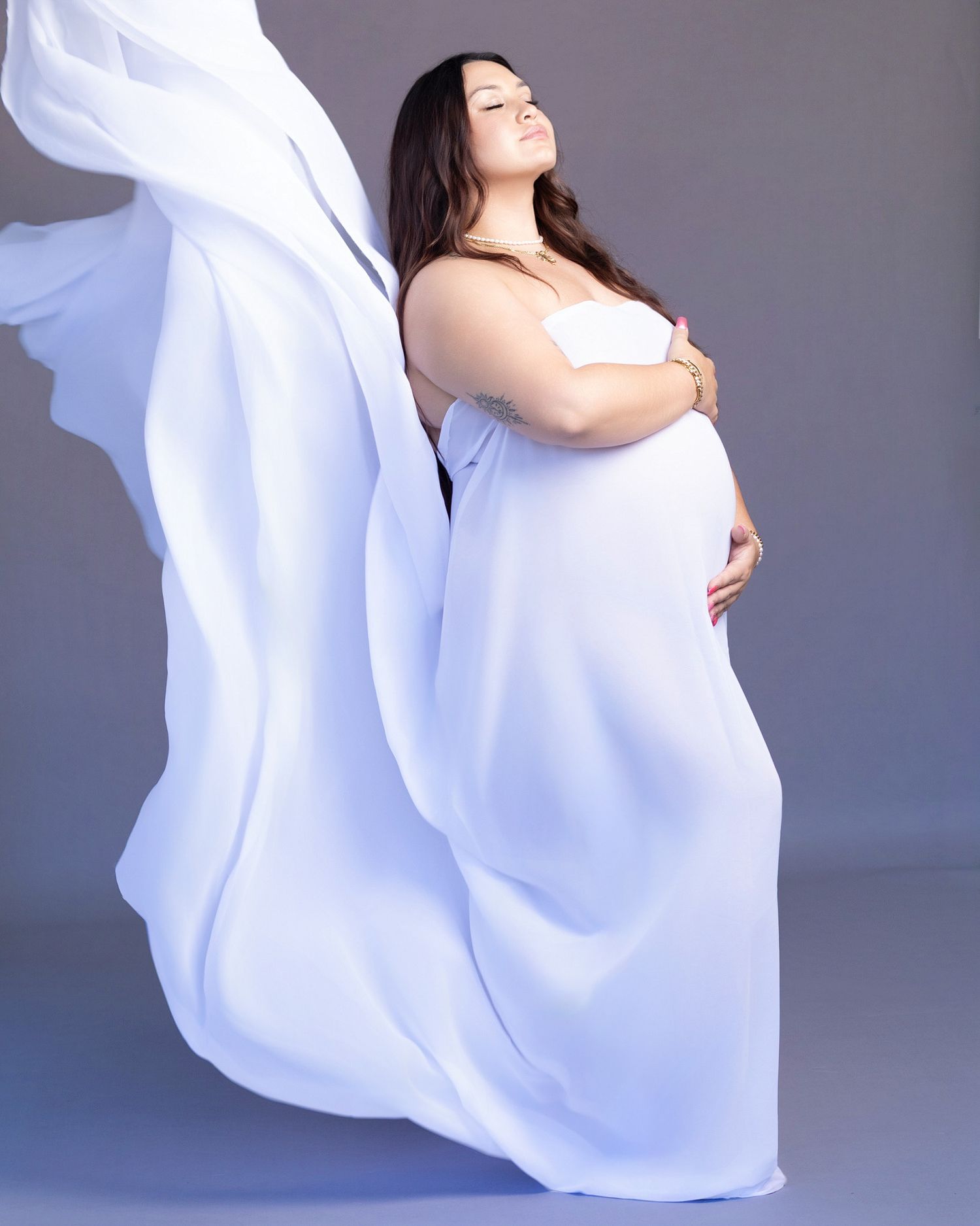 A pregnant woman poses gracefully in a flowing white fabric dress billowing in the air during a studio maternity shoot.