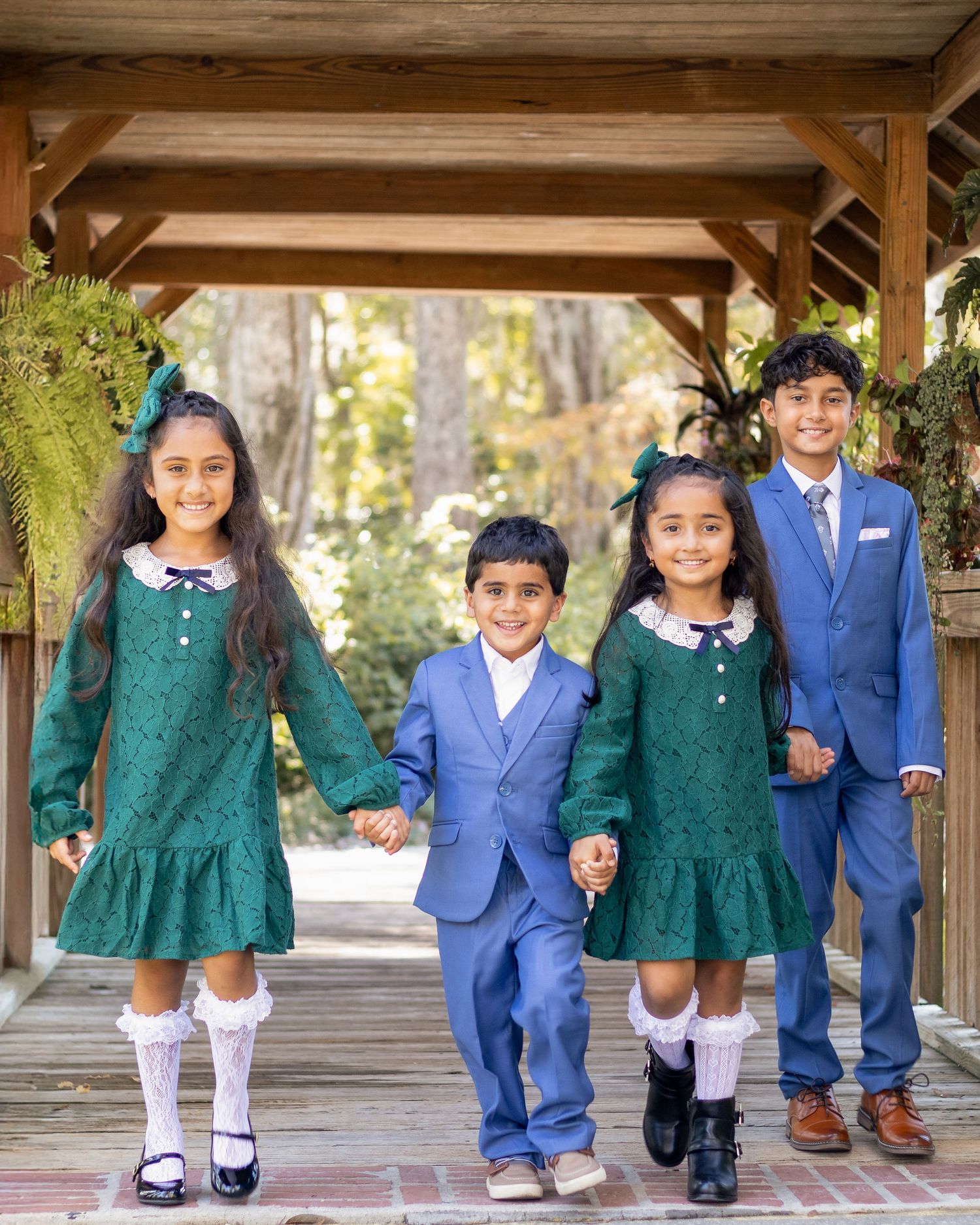 Four children in formal attire, two girls in green dresses and two boys in blue suits, walking hand in hand under a wooden pergola.