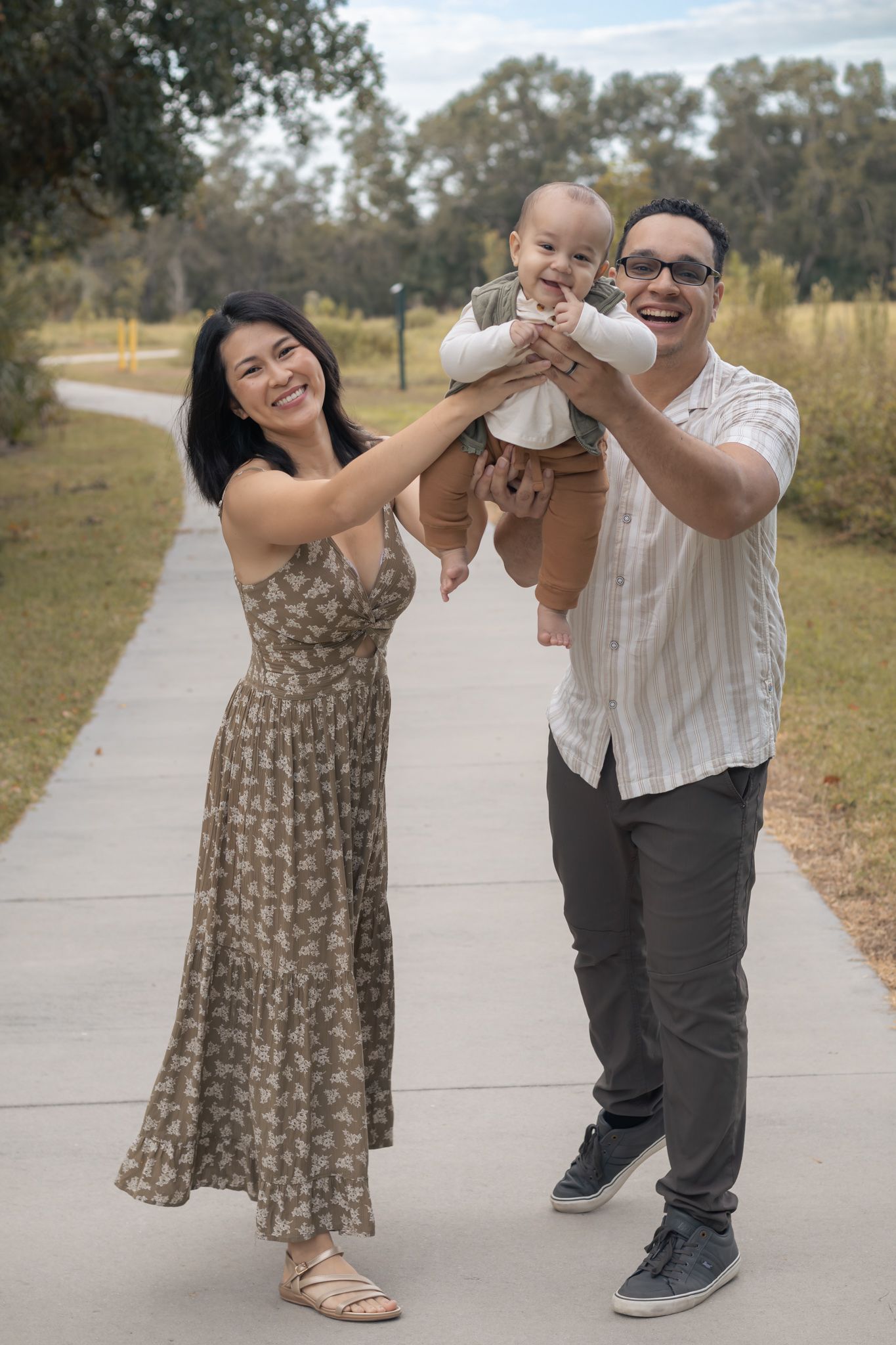 Happy family with mother in floral dress and father lifting smiling baby on a sunny park pathway.