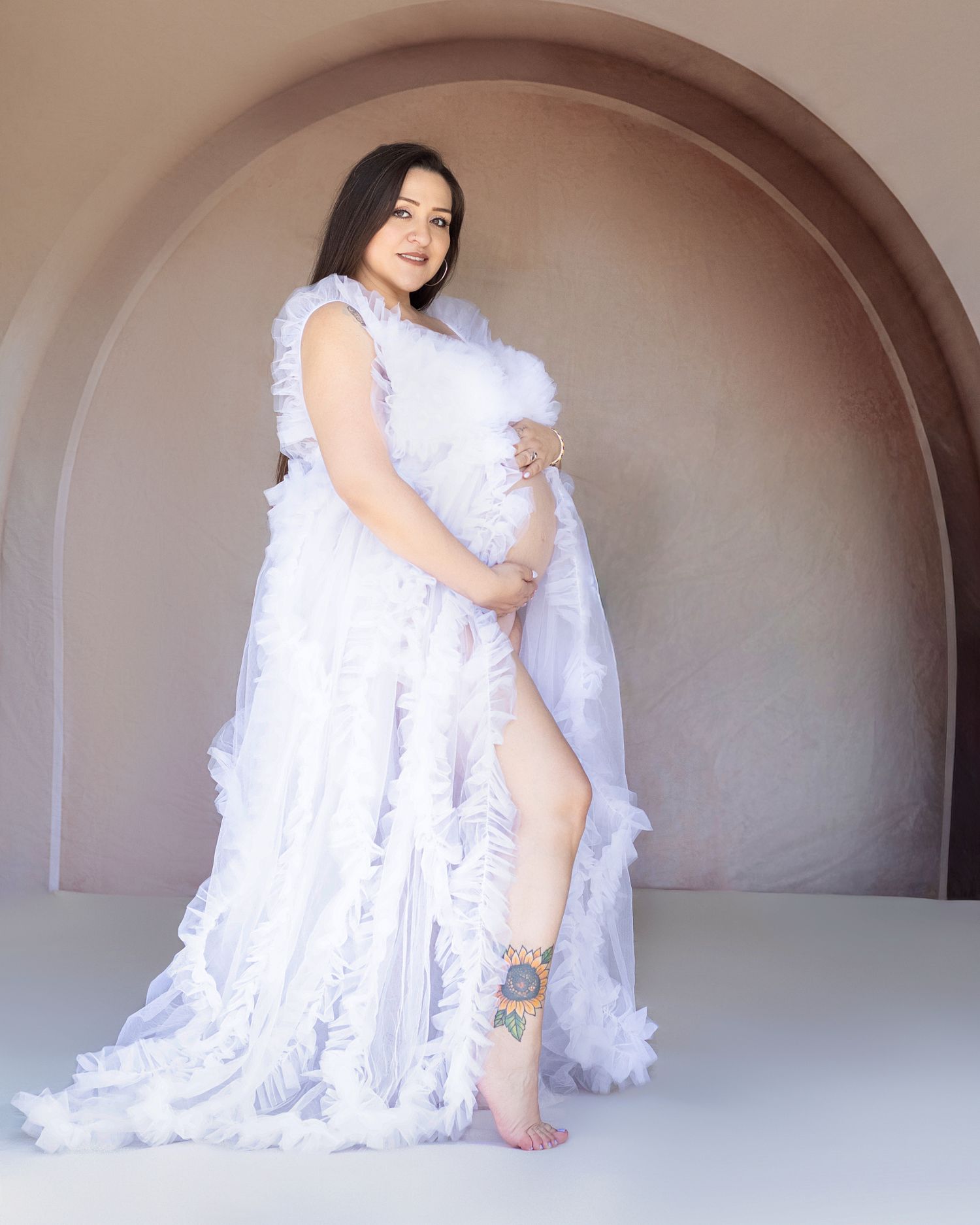Pregnant woman in white ruffled maternity gown posing under arch, showcasing elegant floral ankle tattoo.