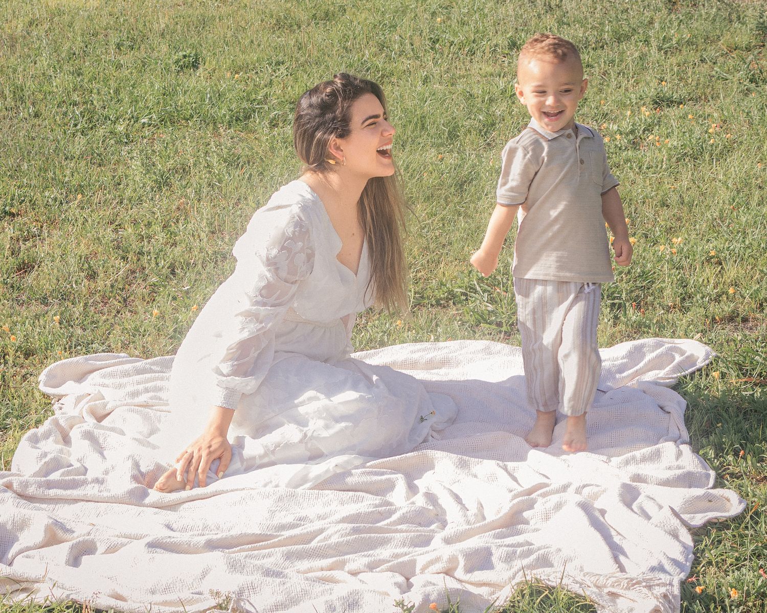 Mother in white dress sitting on blanket in grass, smiling at toddler boy standing beside her.
