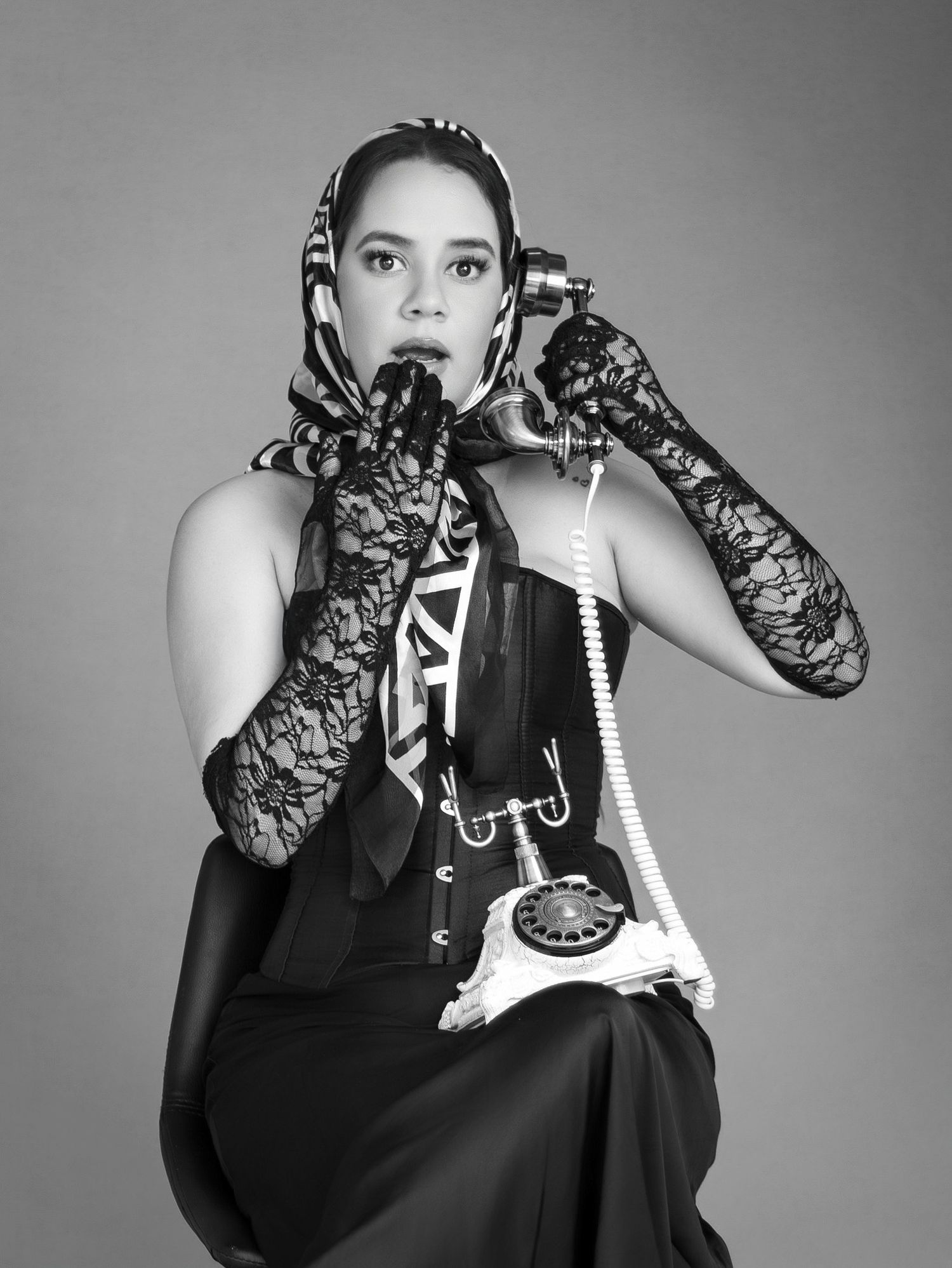 Black and white photo of a woman in lace gloves and headscarf holding a vintage rotary telephone.