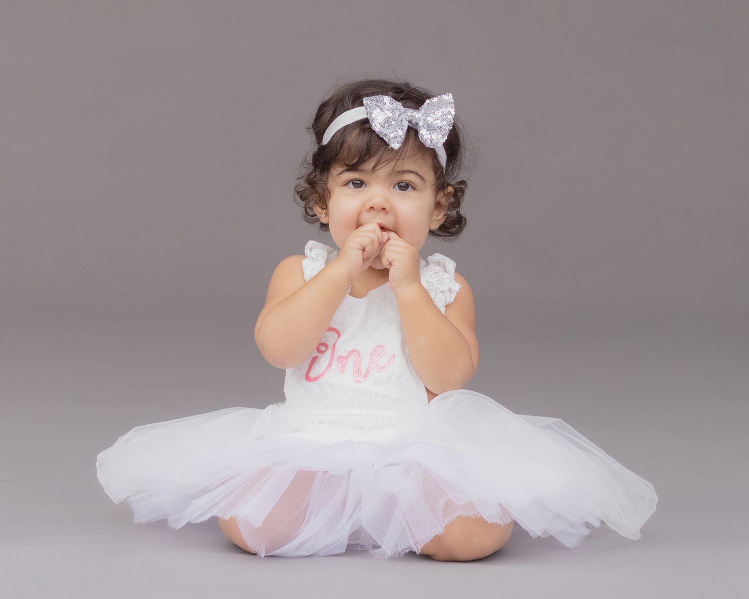 Adorable baby girl in white tutu dress and bow headband sitting on gray background celebrating her first birthday.