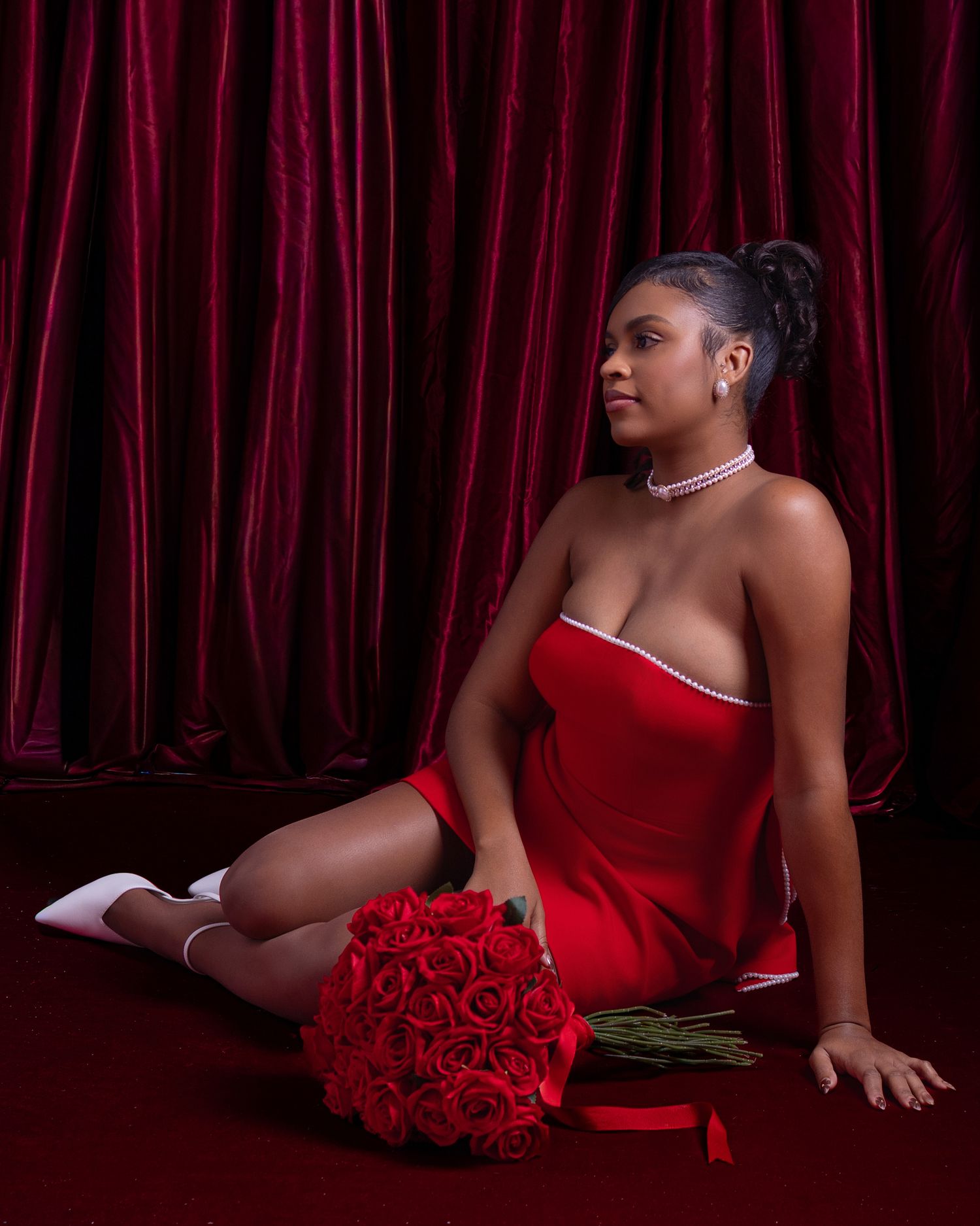 Woman in red strapless dress sitting on floor with rose bouquet against red curtains.