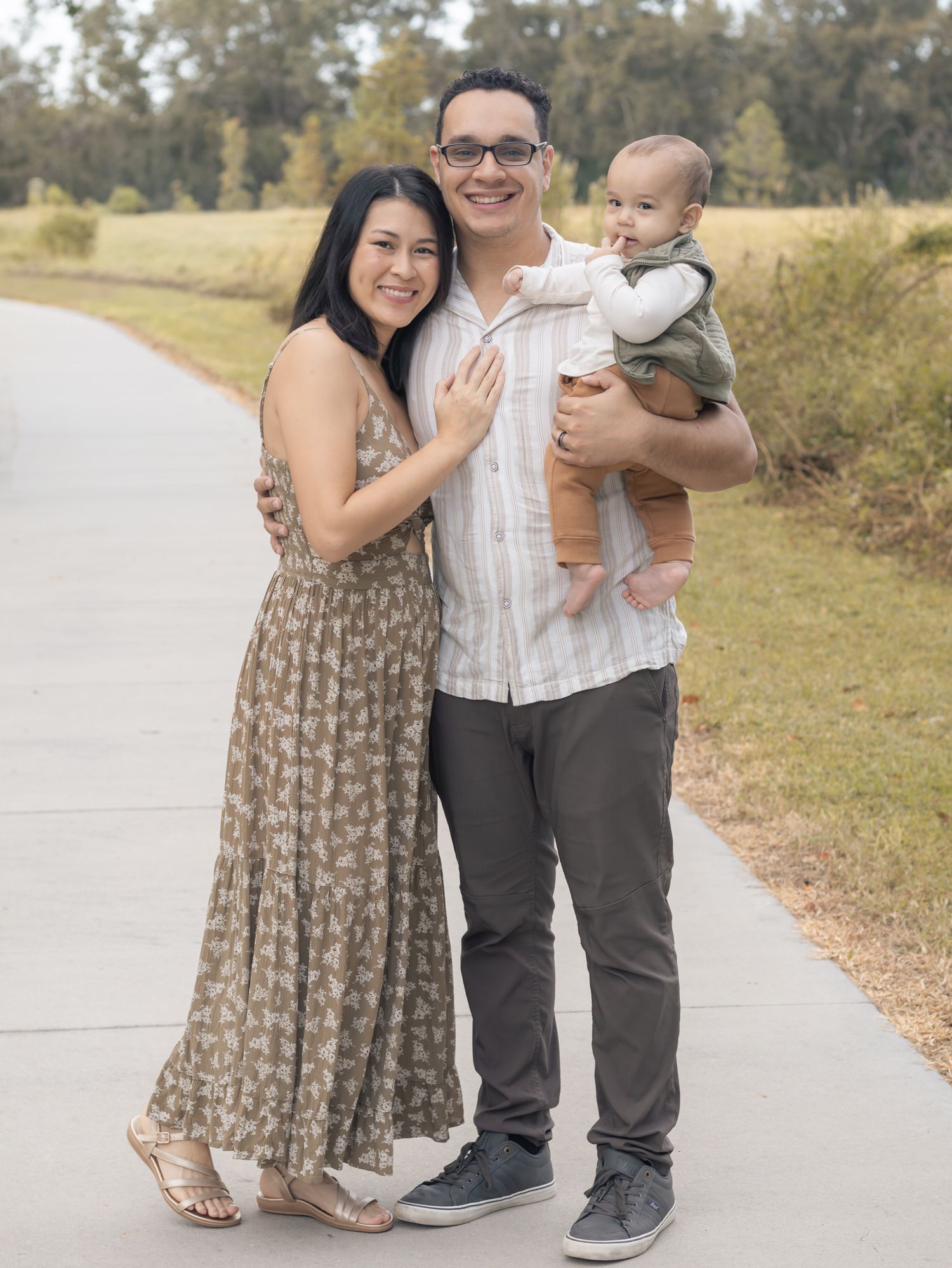 Happy young family with mother, father, and baby posing on a scenic outdoor pathway surrounded by greenery.