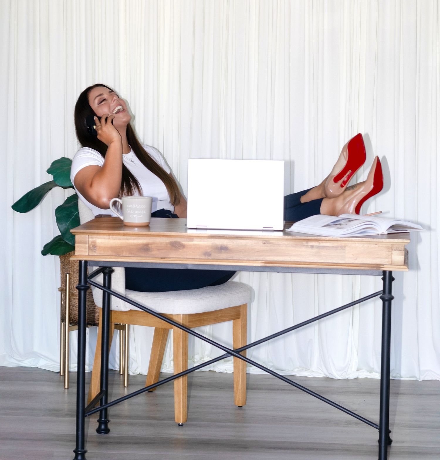 Woman relaxing at wooden desk with laptop, talking on phone with red heels up.