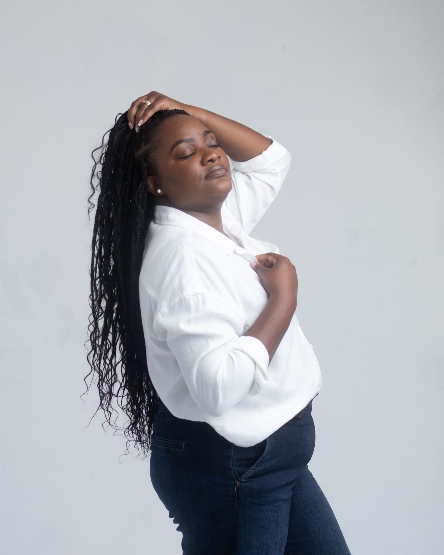 A young Black woman in a white button-up shirt and jeans poses gracefully with hand in hair against a light gray background.