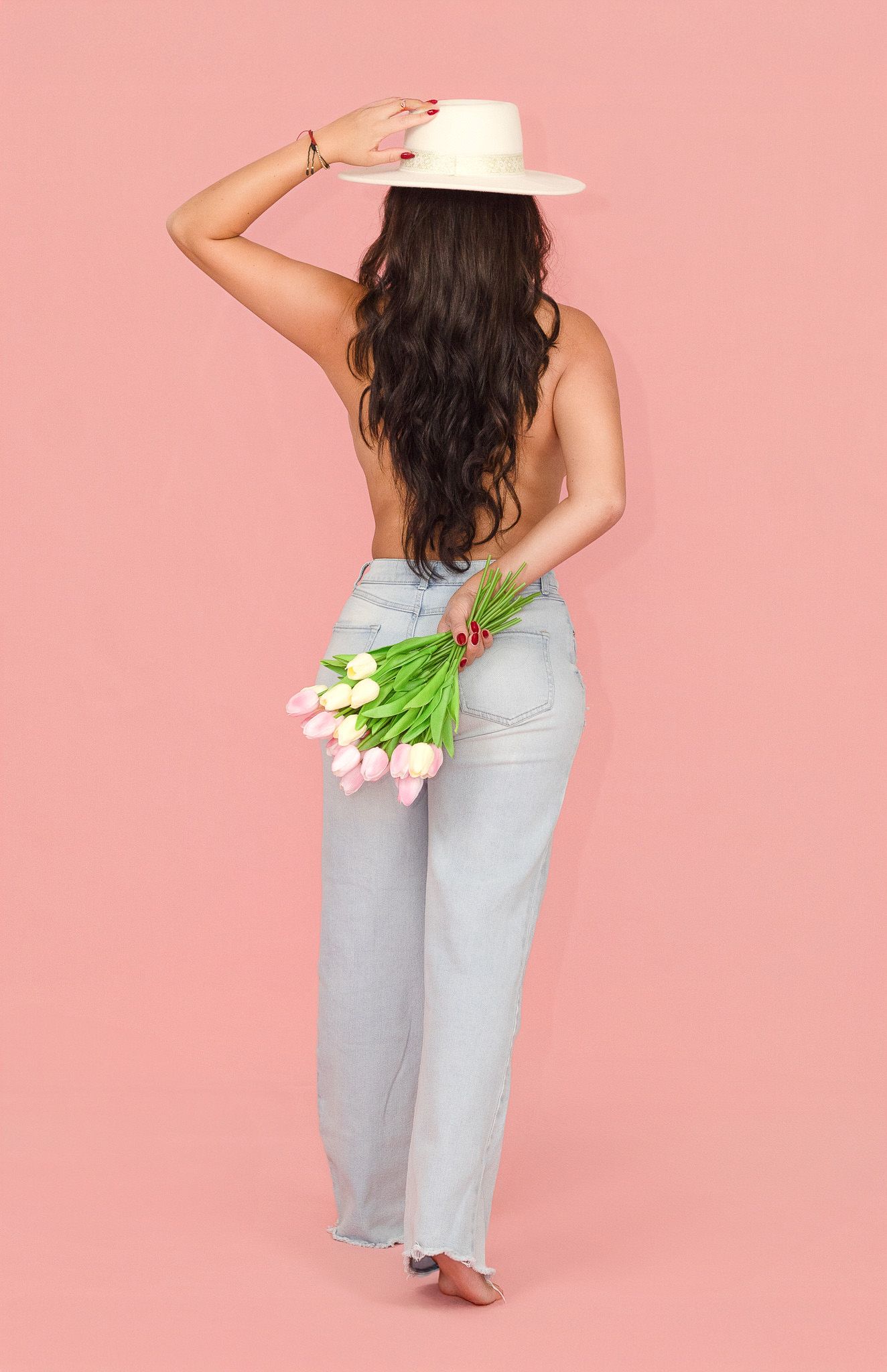 Woman with long dark hair wearing a white hat and light wash jeans, holding pink tulips behind her back on a pink background.