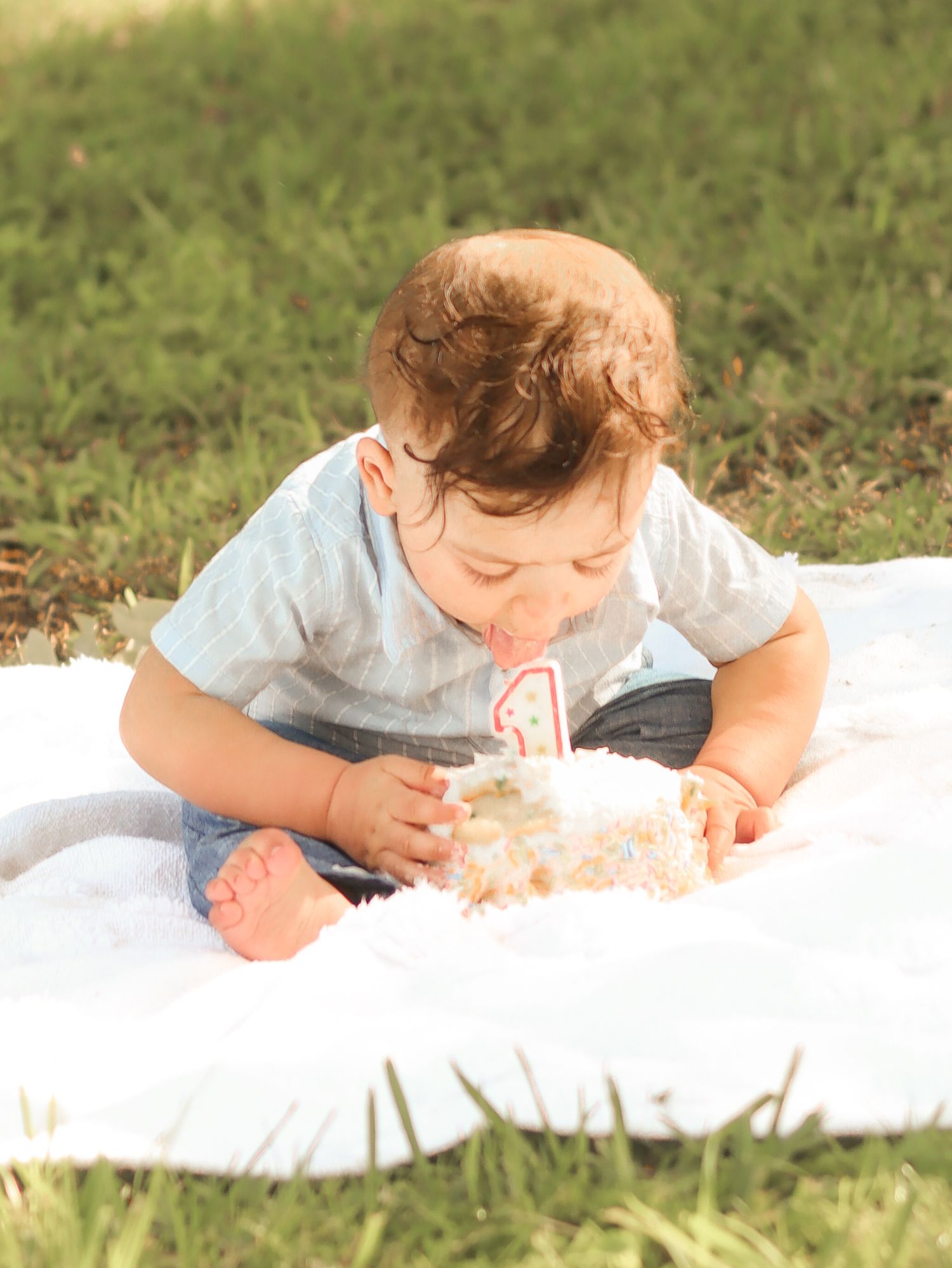 Baby boy leaning into first birthday smash cake on white blanket in grassy park.