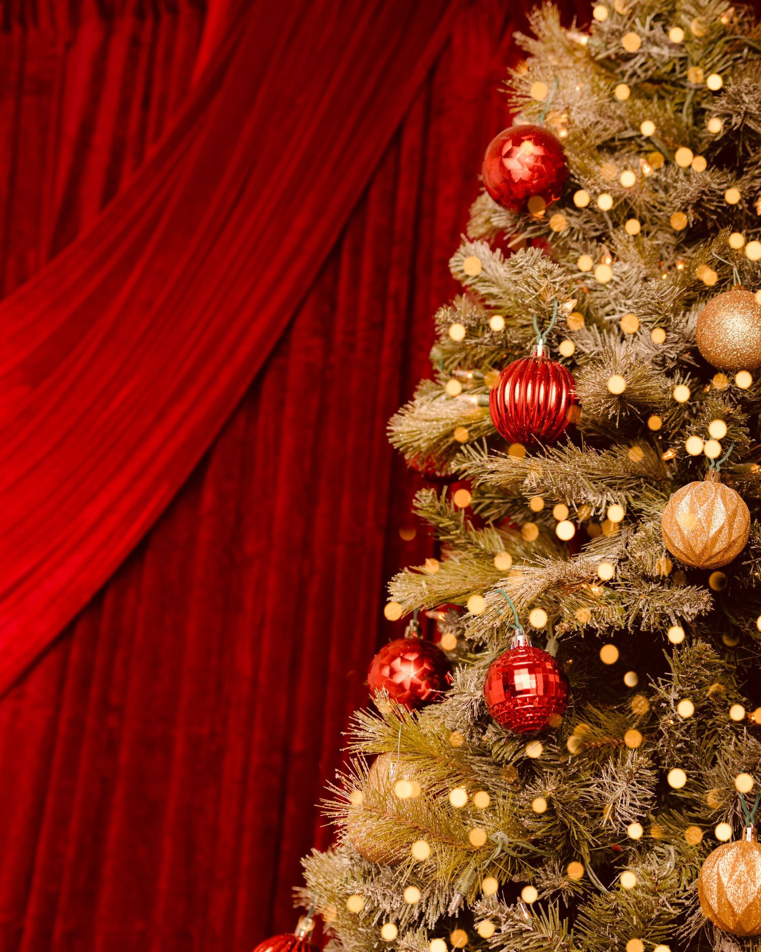 A decorated Christmas tree with red and gold ornaments and twinkling lights beside elegant red velvet curtains.