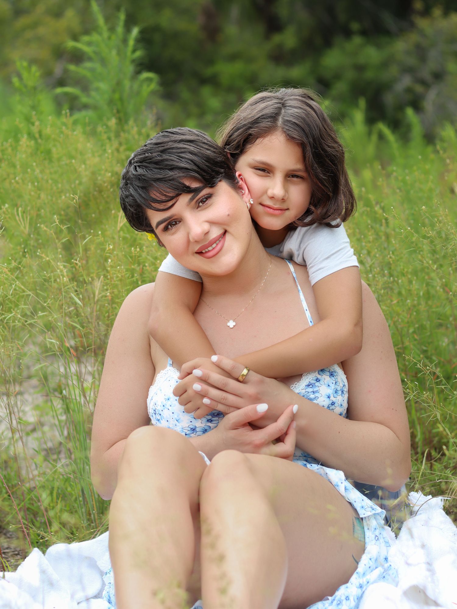 A smiling mother and daughter share a warm embrace while sitting outdoors surrounded by lush green foliage.