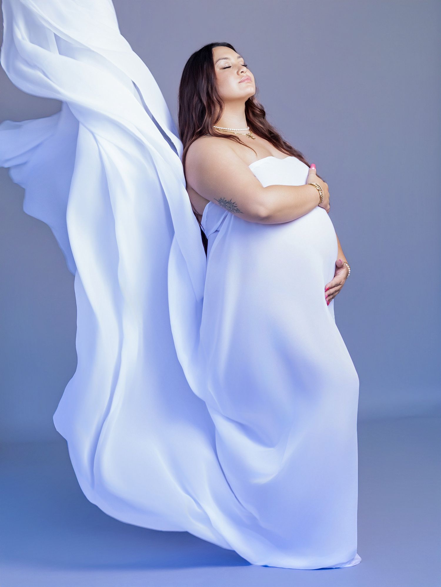 Pregnant woman in flowing white fabric dress posing gracefully against a soft blue-gray studio background.