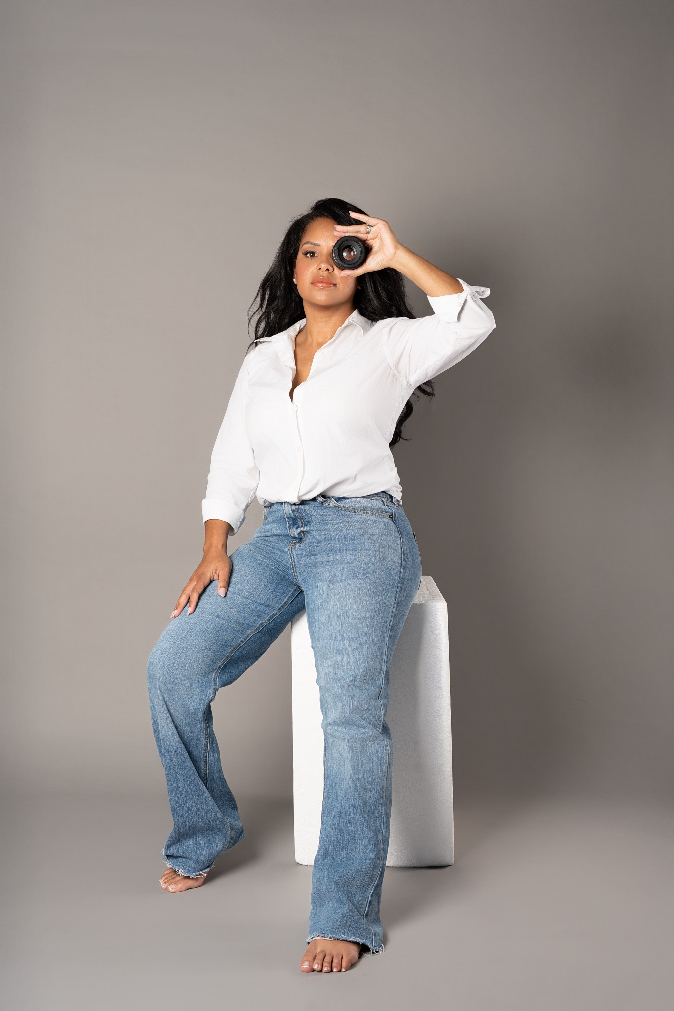 Woman in white shirt and jeans sitting on white cube holding camera to eye.
