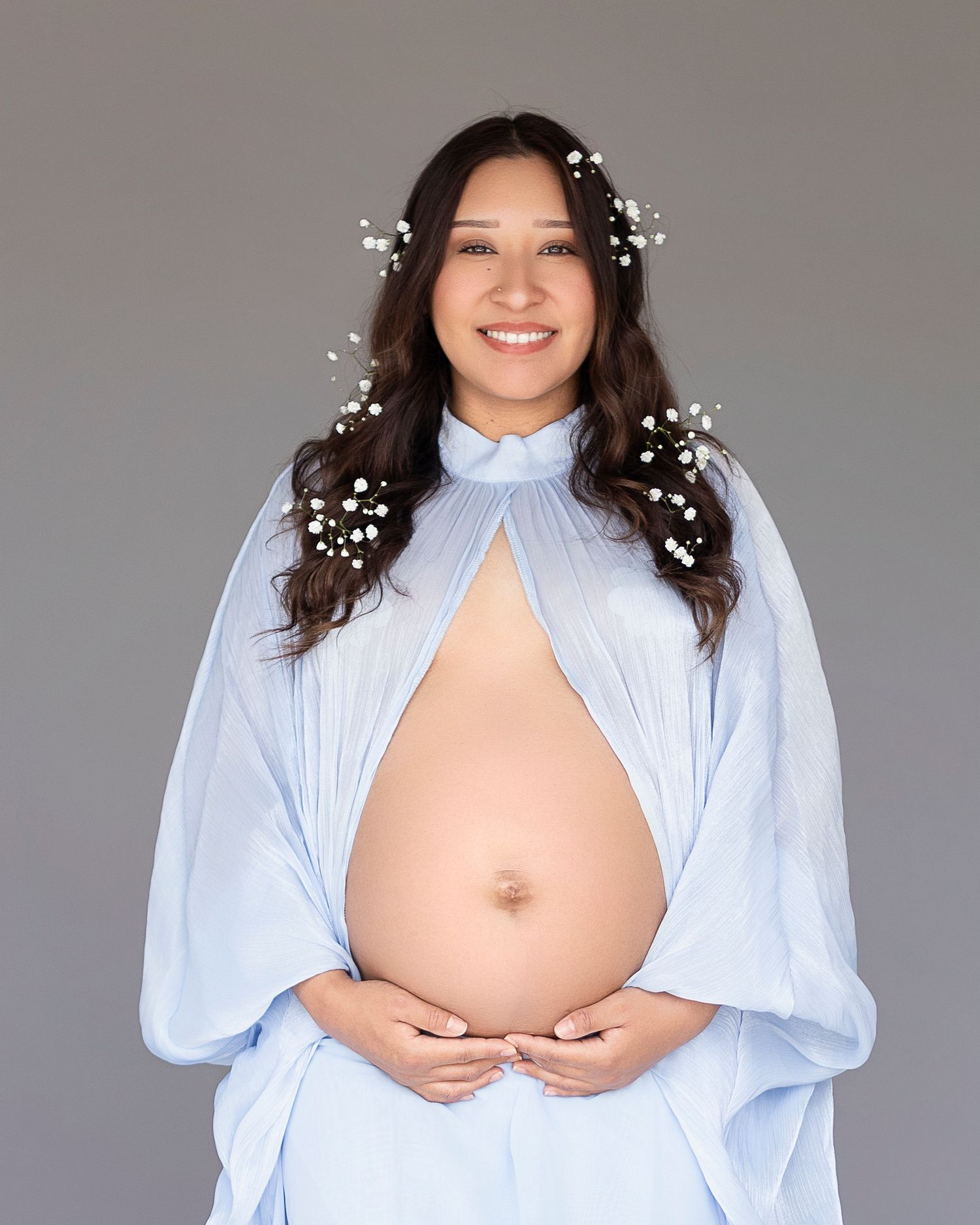 Smiling pregnant woman in light blue cape dress with baby's breath flowers in dark hair, cradling bare baby bump.