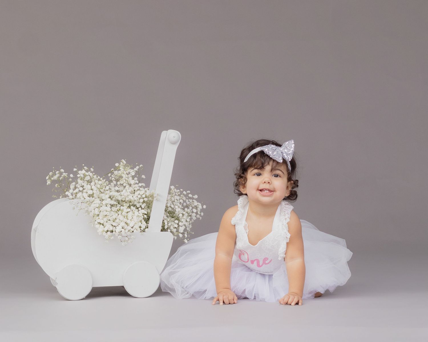 Adorable baby girl in white tutu dress sitting near floral cart on gray background.