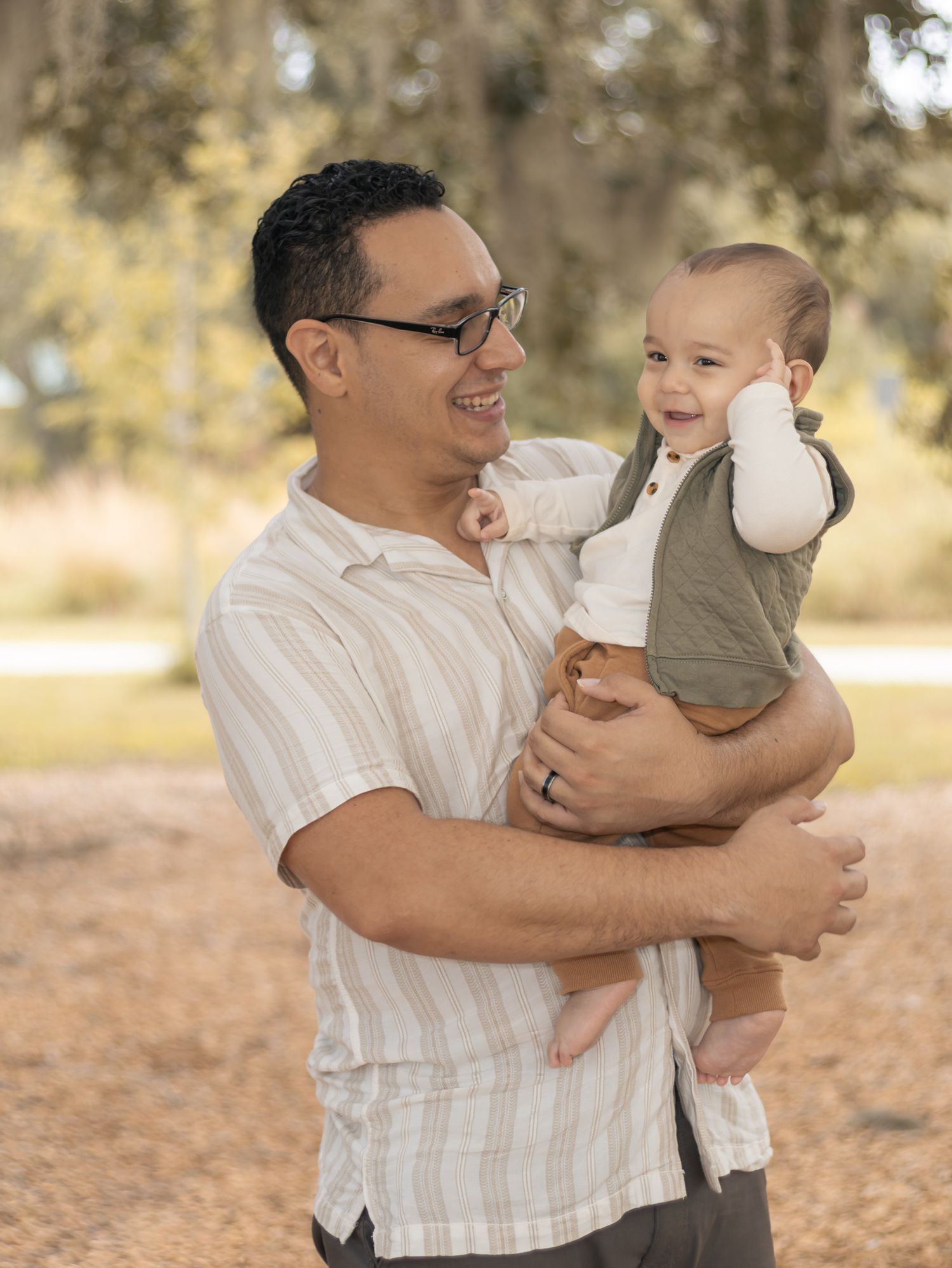 A smiling man with glasses holds a happy baby in a green vest outdoors in a sunny park setting.
