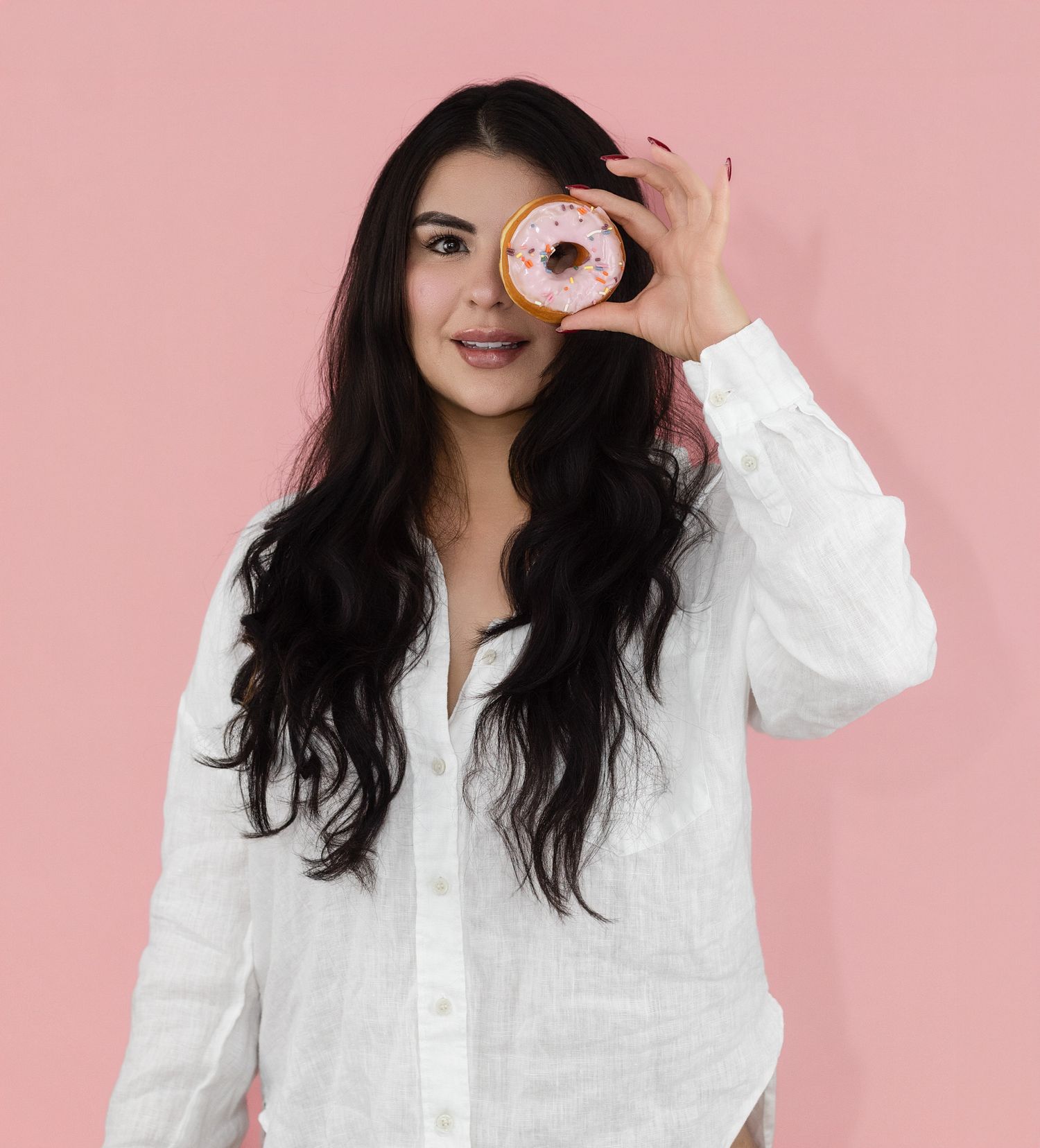 Young woman in white linen shirt holding a pink frosted donut over her eye against a pink background.