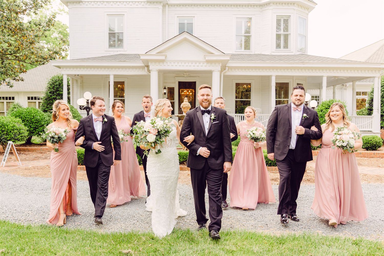 Wedding party walks and poses together outside a classic white mansion.