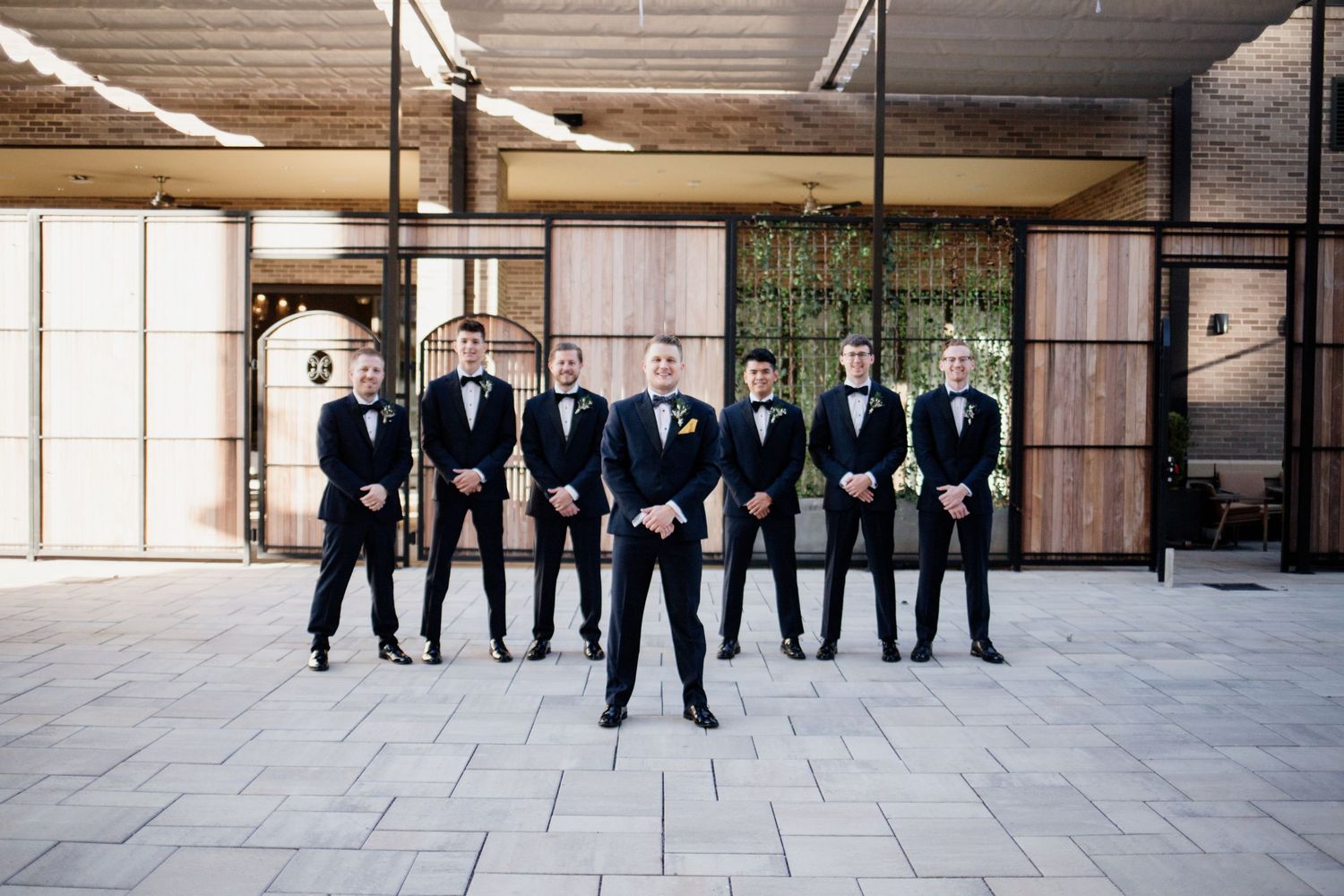 Groomsmen in black tuxedos pose together in industrial indoor space.