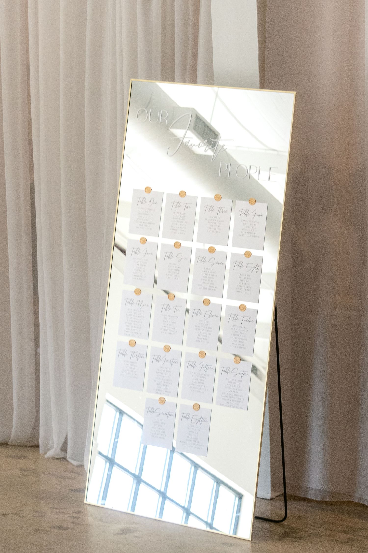 A large full-length mirror displays a wedding seating chart with white papers arranged in neat rows against a light curtain background.