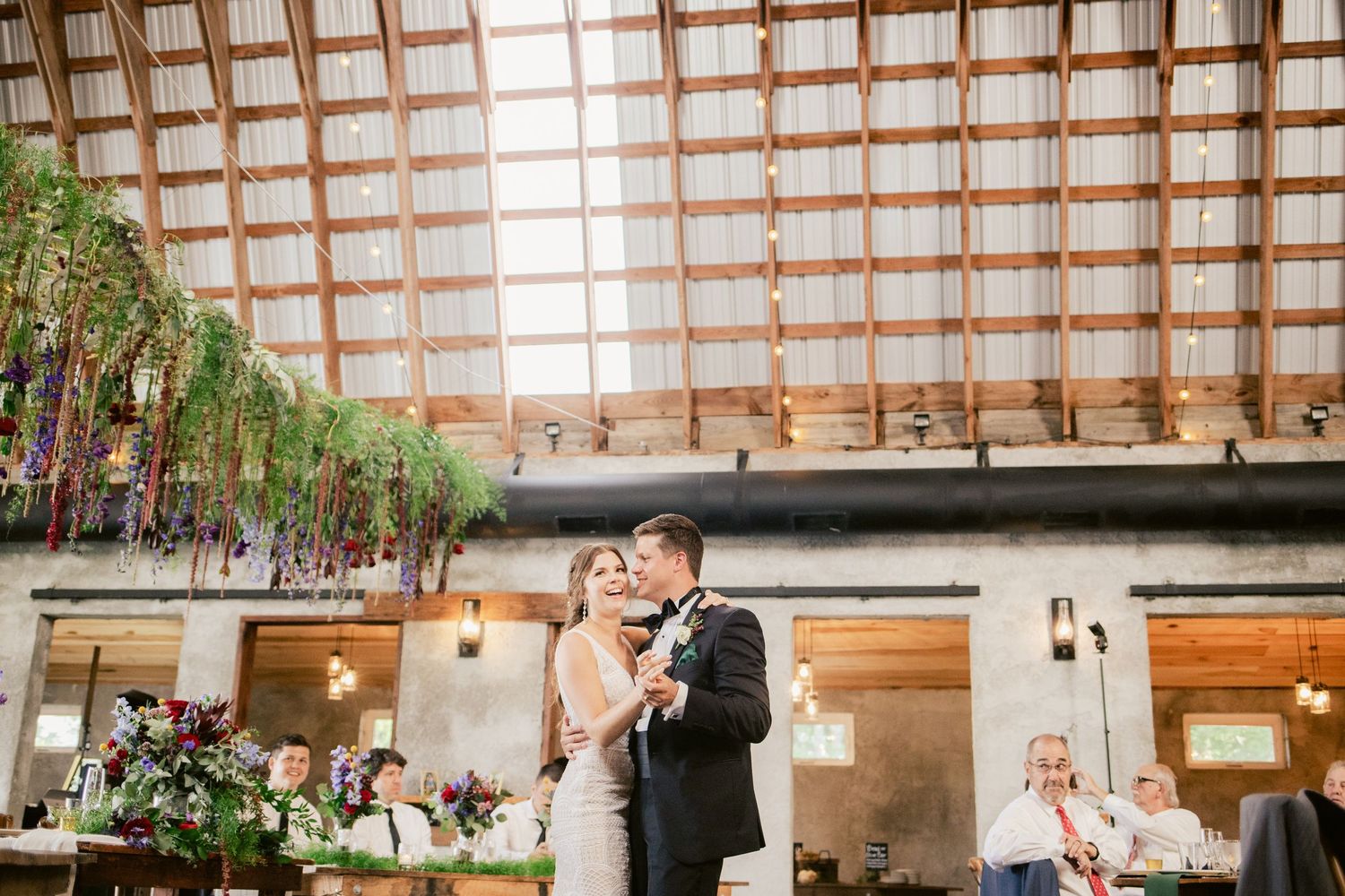 A couple dancing beneath wooden beams and hanging floral decorations at their wedding venue.